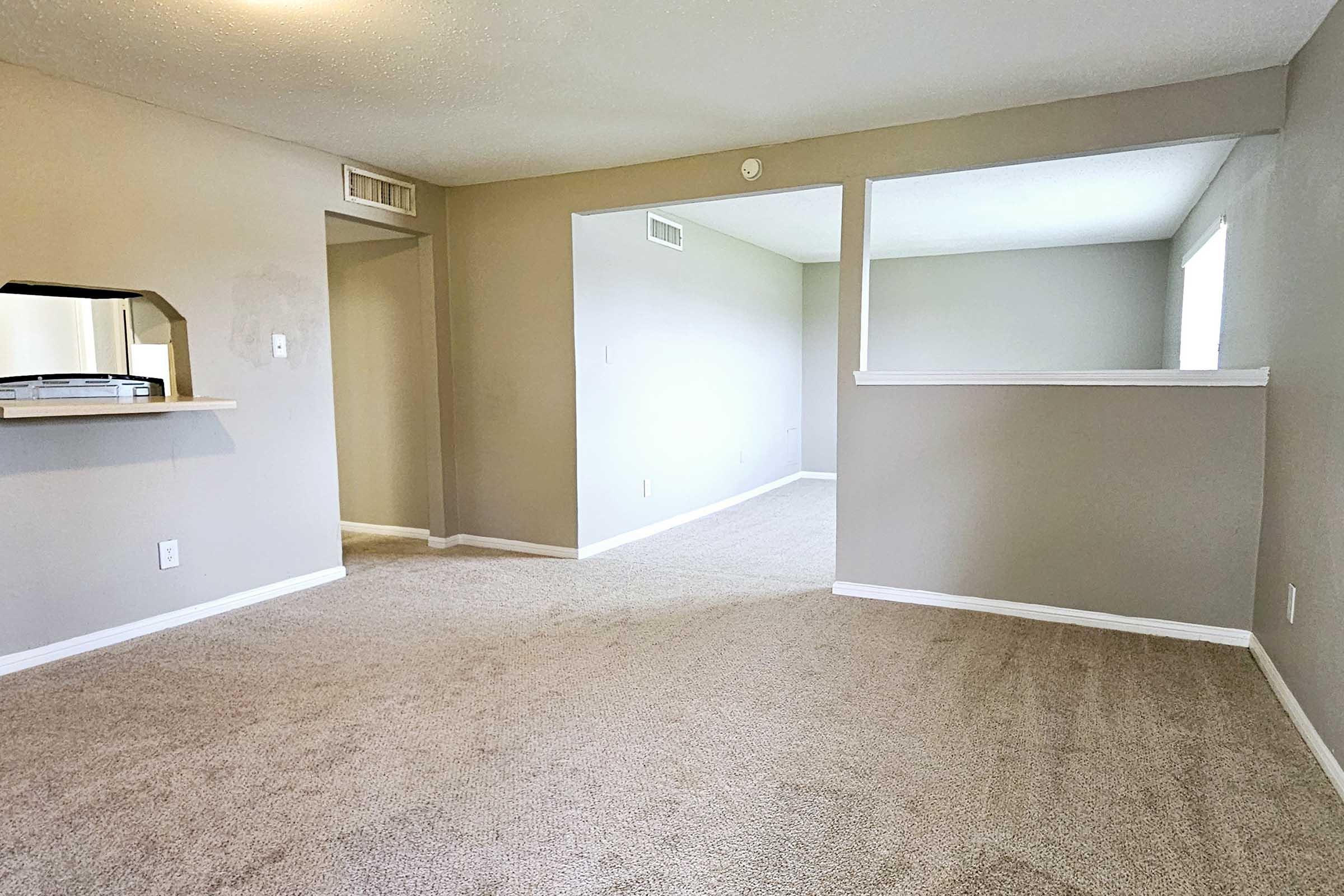 A spacious, carpeted living area with neutral-colored walls. The room features an open layout with a doorway leading to another area. Natural light fills the space from a nearby window, and there is a simple countertop partition viewed from the living space.