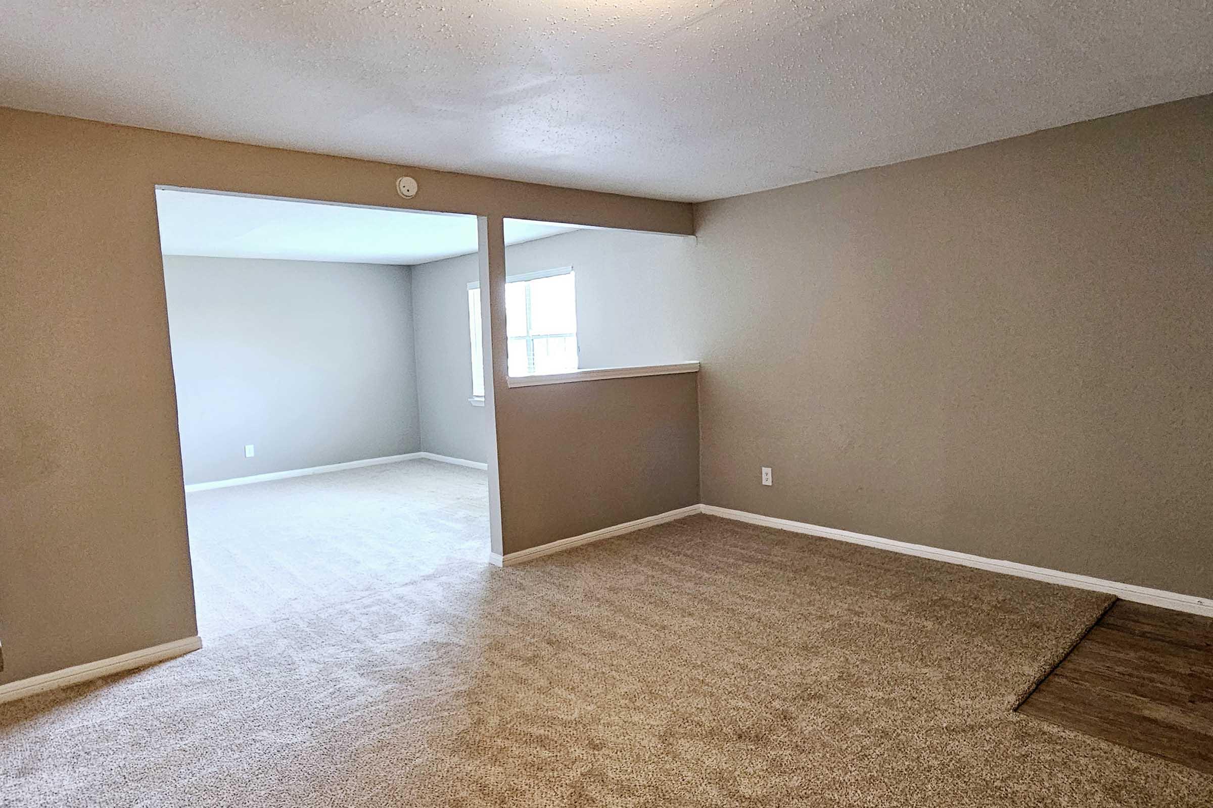 A spacious, light-filled living area featuring neutral-colored walls and carpeted floors. The room has an open layout with a partial wall dividing it from another space, which is empty and also well-lit by natural light coming through a window.