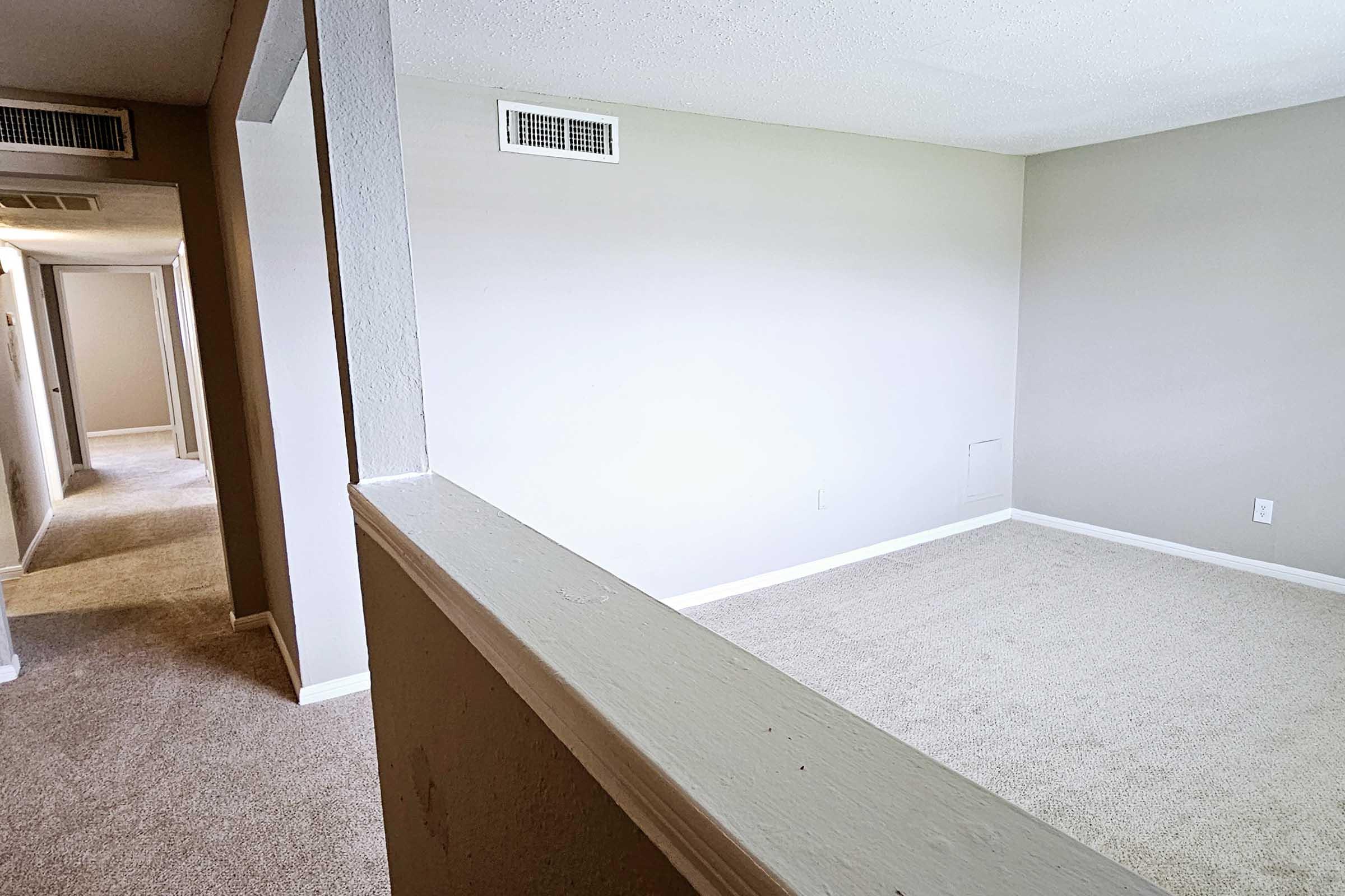 A wide-angle view of a vacant room with light-colored walls and carpet. A hallway is visible on the left, leading to other doors. The room is bright and spacious, with a simple design, and a small wall opening in the corner reveals a concealed area.