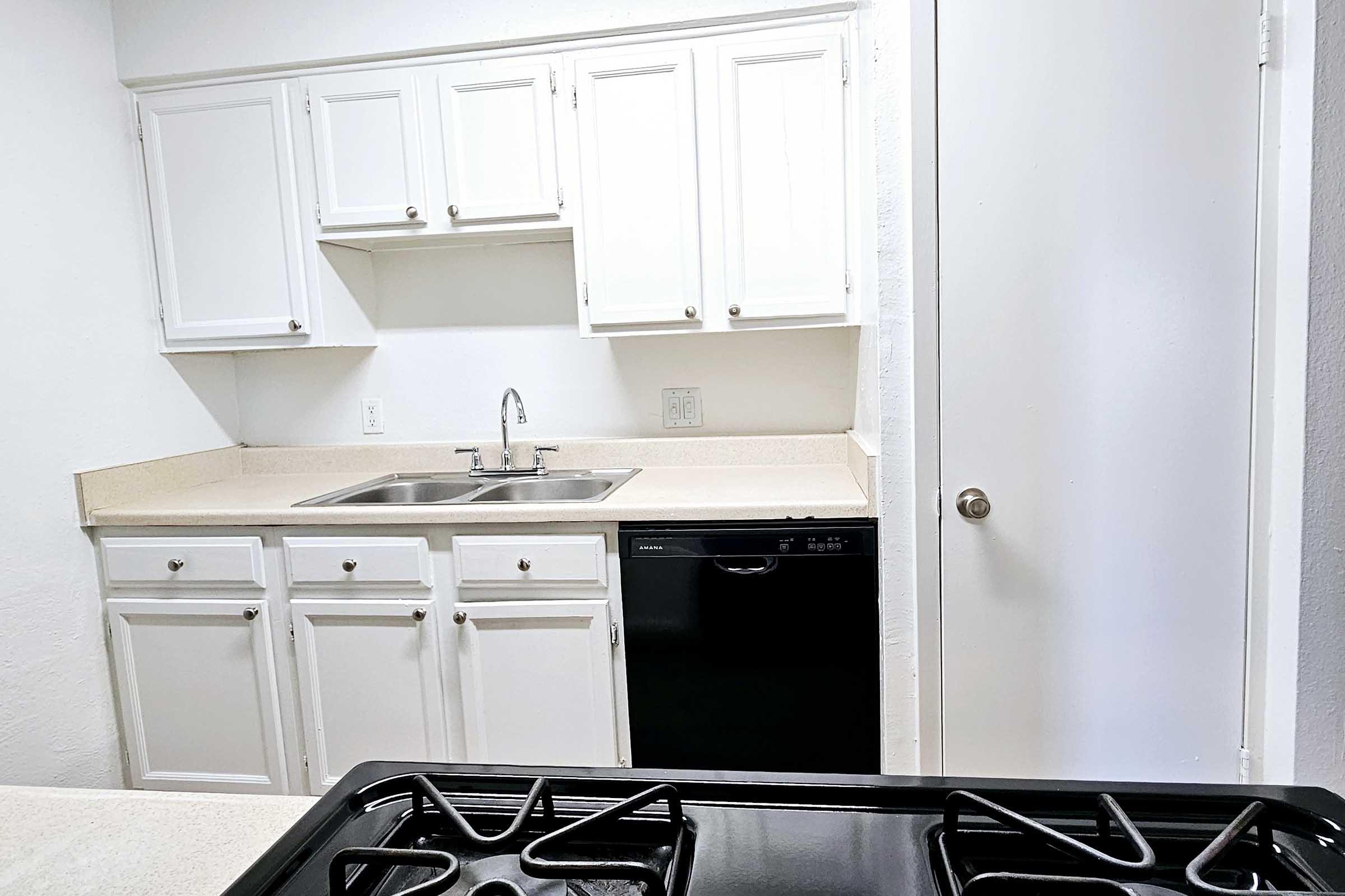 A modern kitchen featuring white cabinets, a double sink, and a black dishwasher. The countertop is light-colored and clean, with a stove visible in the foreground. A closed door is seen on the right, and the overall space appears bright and minimalistic.