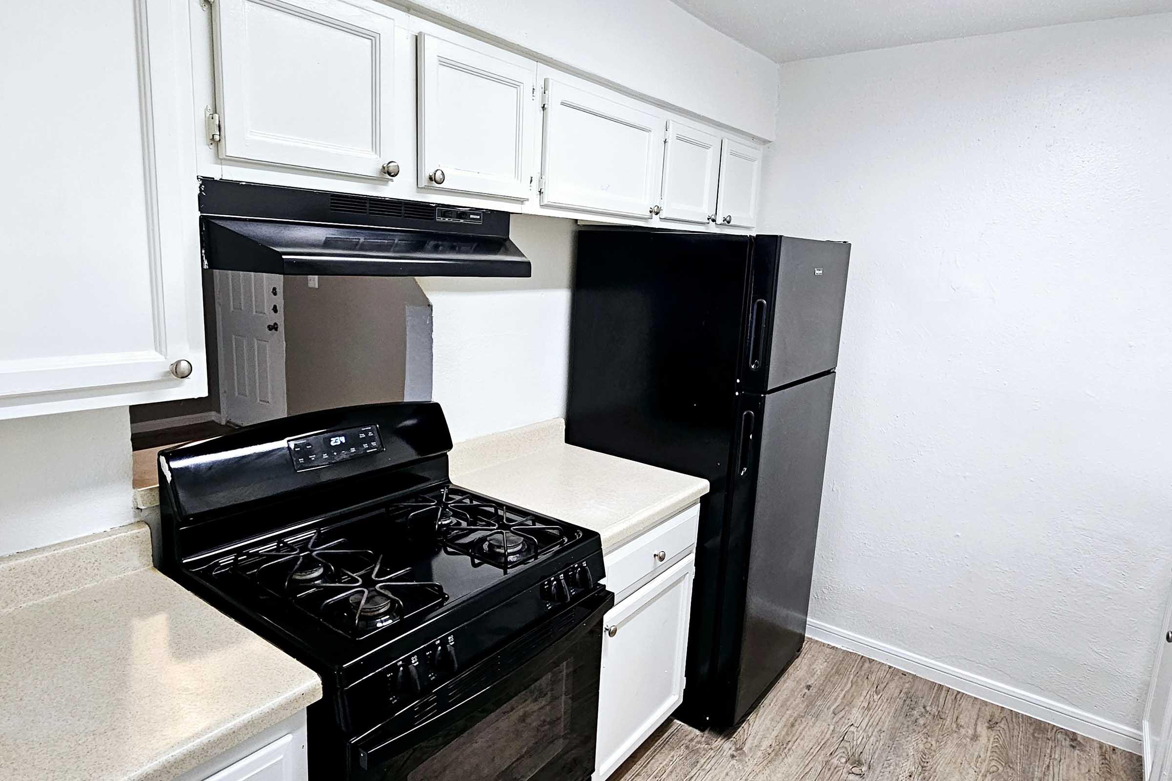 A kitchen featuring white cabinetry, a black gas stove, an over-the-range microwave, and a black refrigerator. The countertop is light-colored, and the floor has a wood-like appearance. The walls are painted white, creating a clean and modern look.