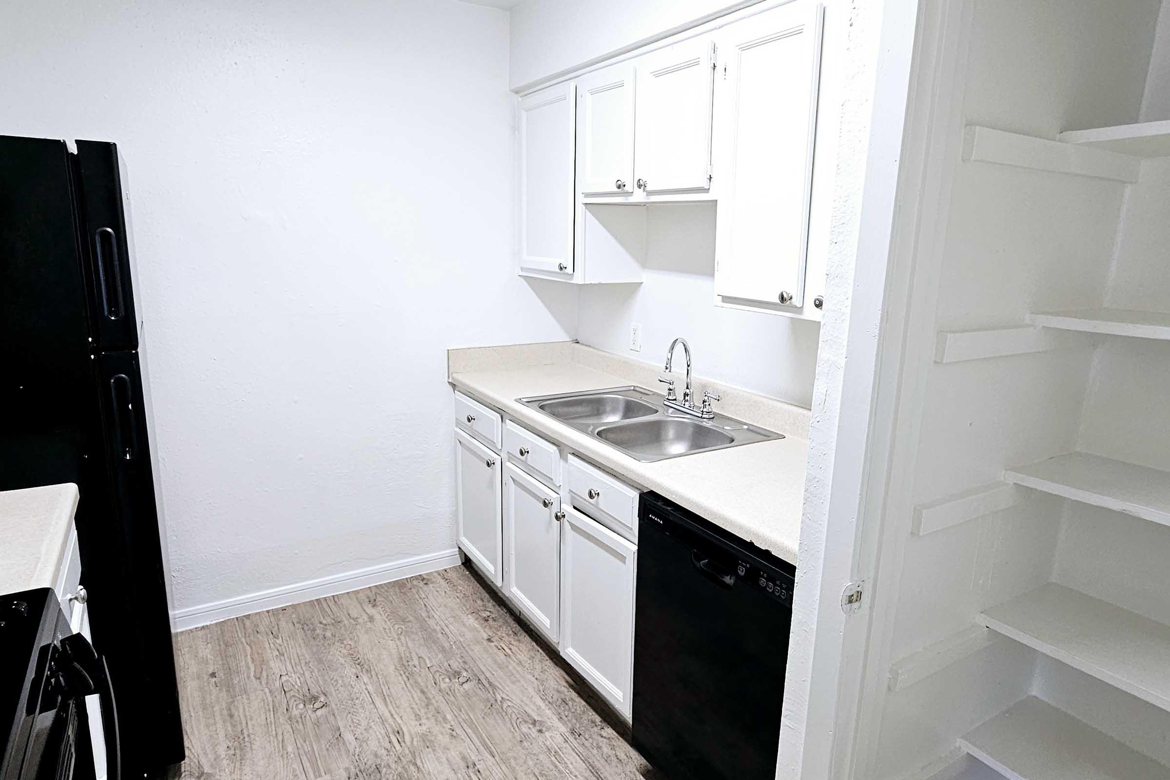 A small, modern kitchen featuring white cabinets, a double sink, and a black dishwasher. The countertops are light-colored, and there is a black refrigerator visible on the left. The walls are painted white, and there is a pantry with open shelving to the right. The flooring is light wood laminate.