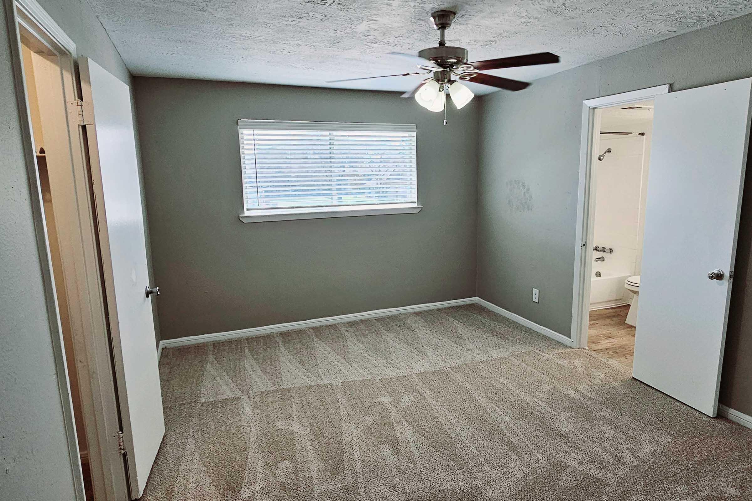 A clean, empty room with a ceiling fan and a large window providing natural light. The walls are painted a neutral gray, and the floor is carpeted. Two doors are visible, one leading to a closet and the other to a bathroom. The room appears freshly painted and well-maintained.