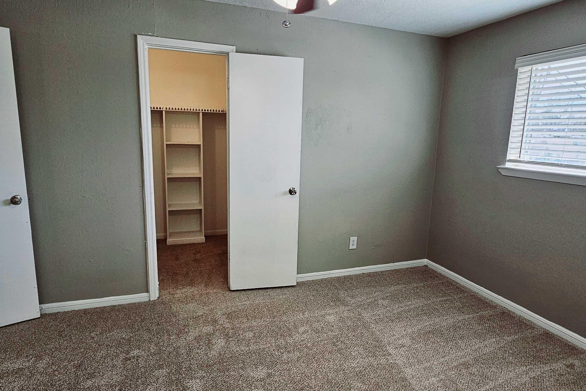 A simple room with gray walls and a carpeted floor. There are two doors, one partially open leading to a small closet with shelves. A window with blinds allows light into the space, creating a bright and airy feel.