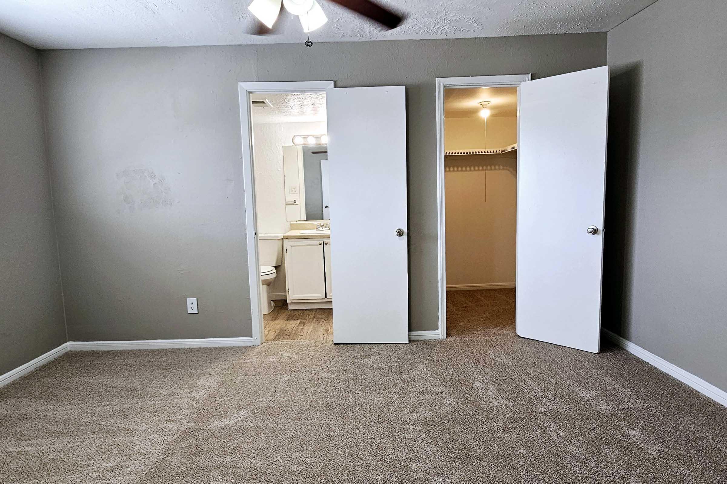 A spacious, neutral-toned bedroom featuring light gray walls and soft carpet. Two white doors lead to a bathroom and a closet. A ceiling fan is visible above, and natural light enters through the doorway. The room appears well-maintained and tidy, enhancing its inviting atmosphere.