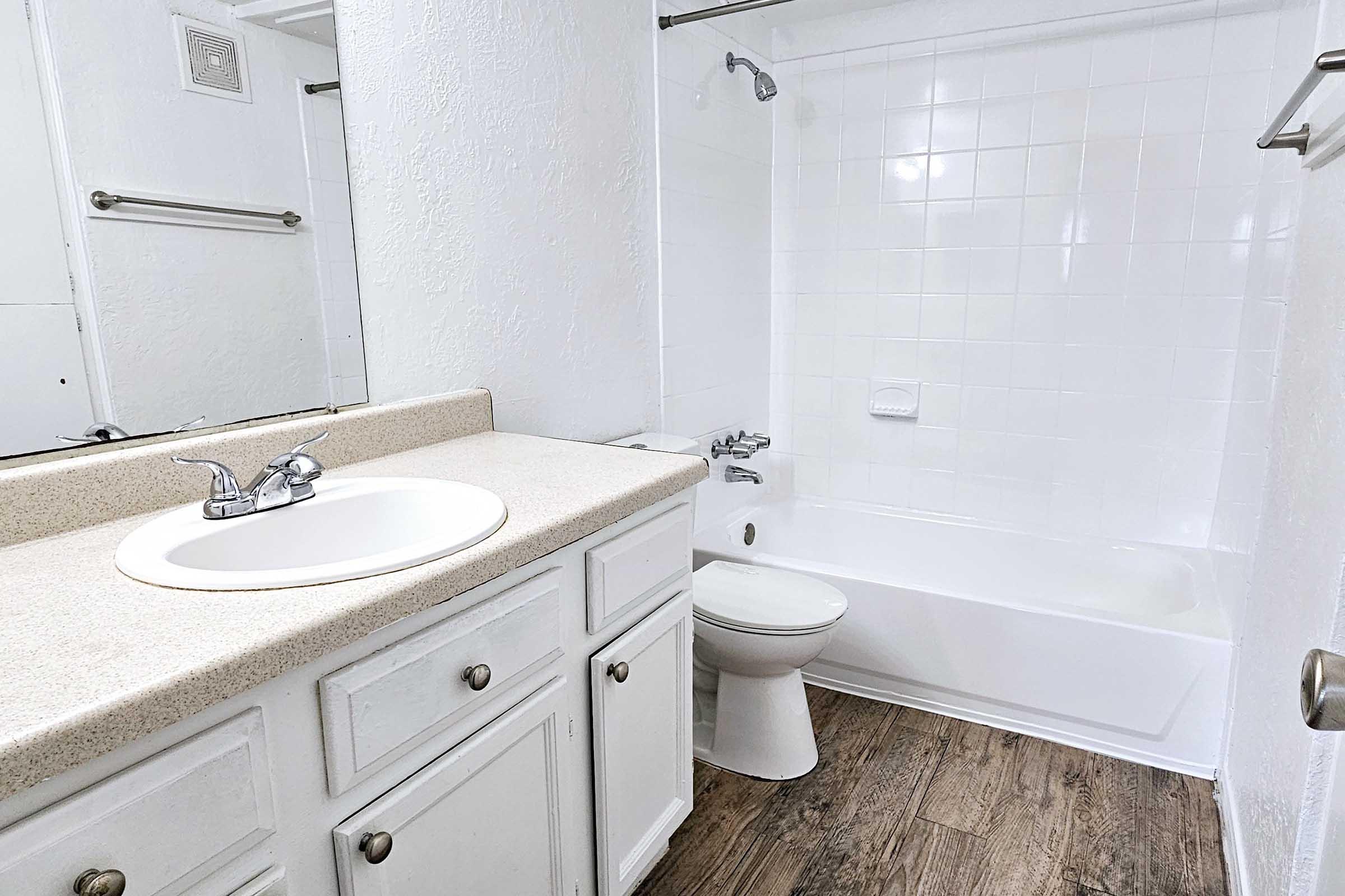 A clean and modern bathroom featuring a white tiled wall, a bathtub with a showerhead, a white toilet, and a vanity with a sink. The floor has a wood-like appearance, and there are mirrors and towel bars on the walls. The overall color scheme is bright and neutral.