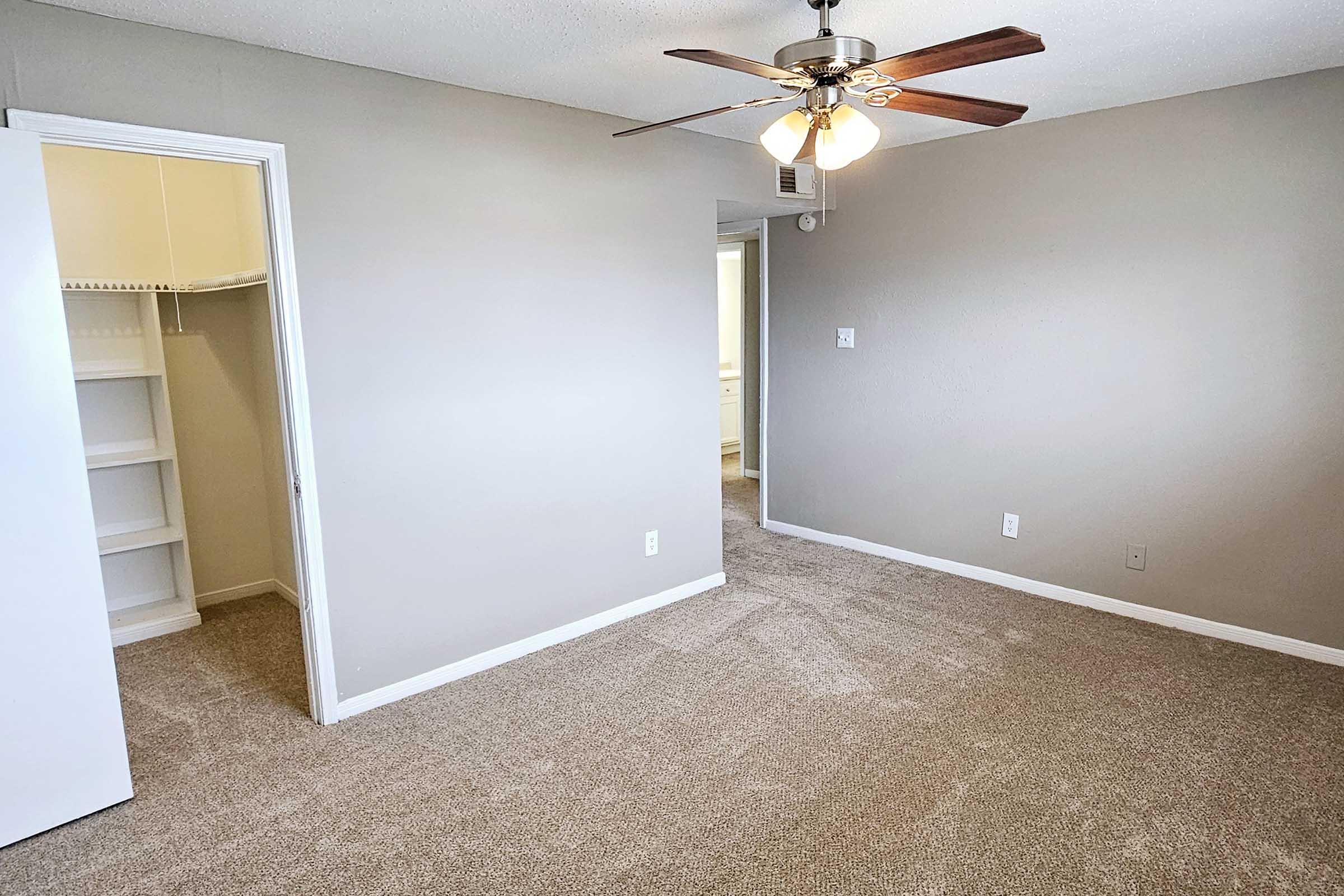 A spacious, empty room with light gray walls and a ceiling fan. The floor is carpeted in a neutral tone. On the left side, there is an open white closet with shelves. A doorway in the back leads to another room or area, suggesting a bright and airy living space.