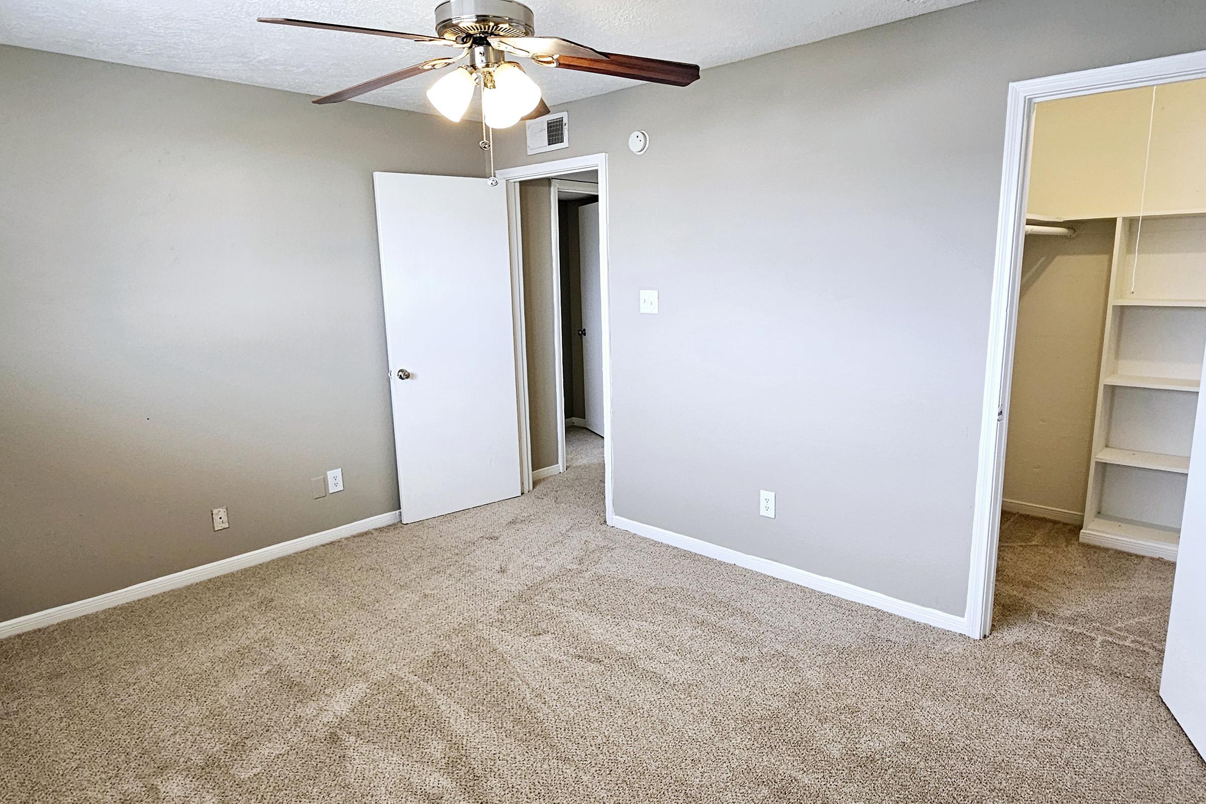 A well-lit, empty room featuring tan carpet and light gray walls. A ceiling fan with four bulbs hangs from the ceiling. One door leads to a closet, while another door exits to a hallway. The room appears clean and ready for furnishings.