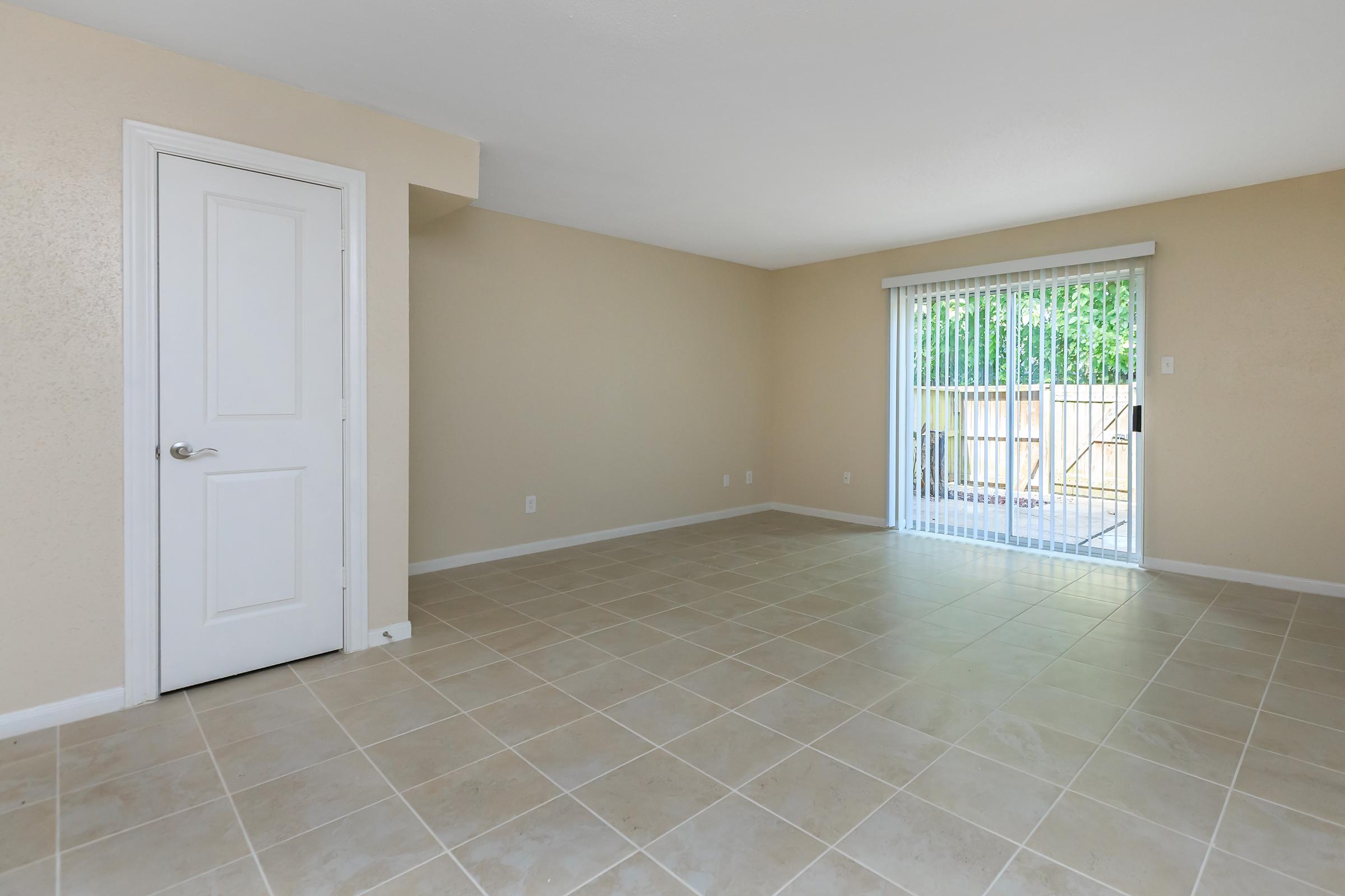 A spacious, empty room with light beige walls and tiled flooring. A white door is visible on the left, and a sliding glass door on the right leads to an outdoor area. The space is well-lit with natural light coming through the door, creating a bright and open atmosphere.