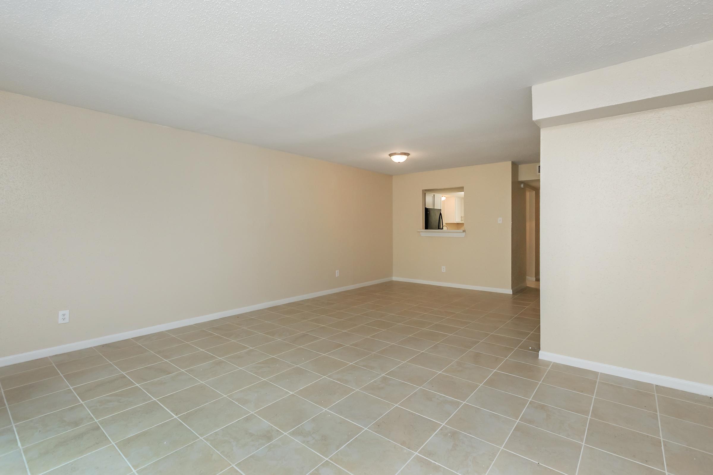 Spacious empty living area with beige walls and tile flooring. A small opening in the wall leads to a kitchenette. The room is well-lit, featuring a ceiling light fixture and a neutral color palette, providing a blank canvas for decorating.