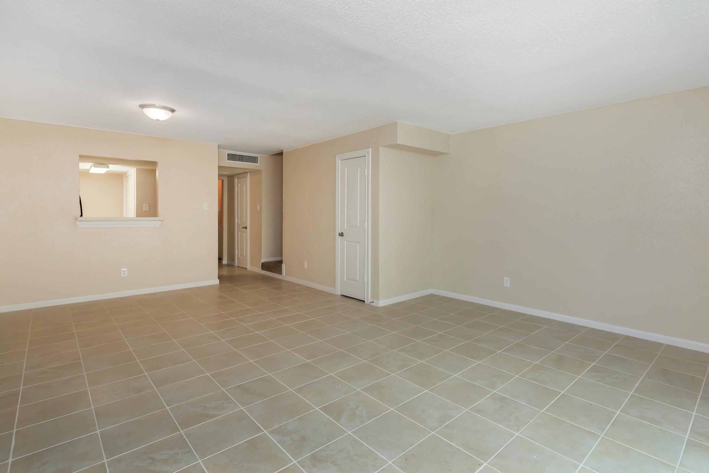 Empty room with beige walls and tiled floor. A light fixture on the ceiling provides illumination. A small opening in the wall suggests a pass-through to a kitchen area, and a door leads to another space out of view. The overall atmosphere is neutral and spacious, suitable for various furnishings and decor.
