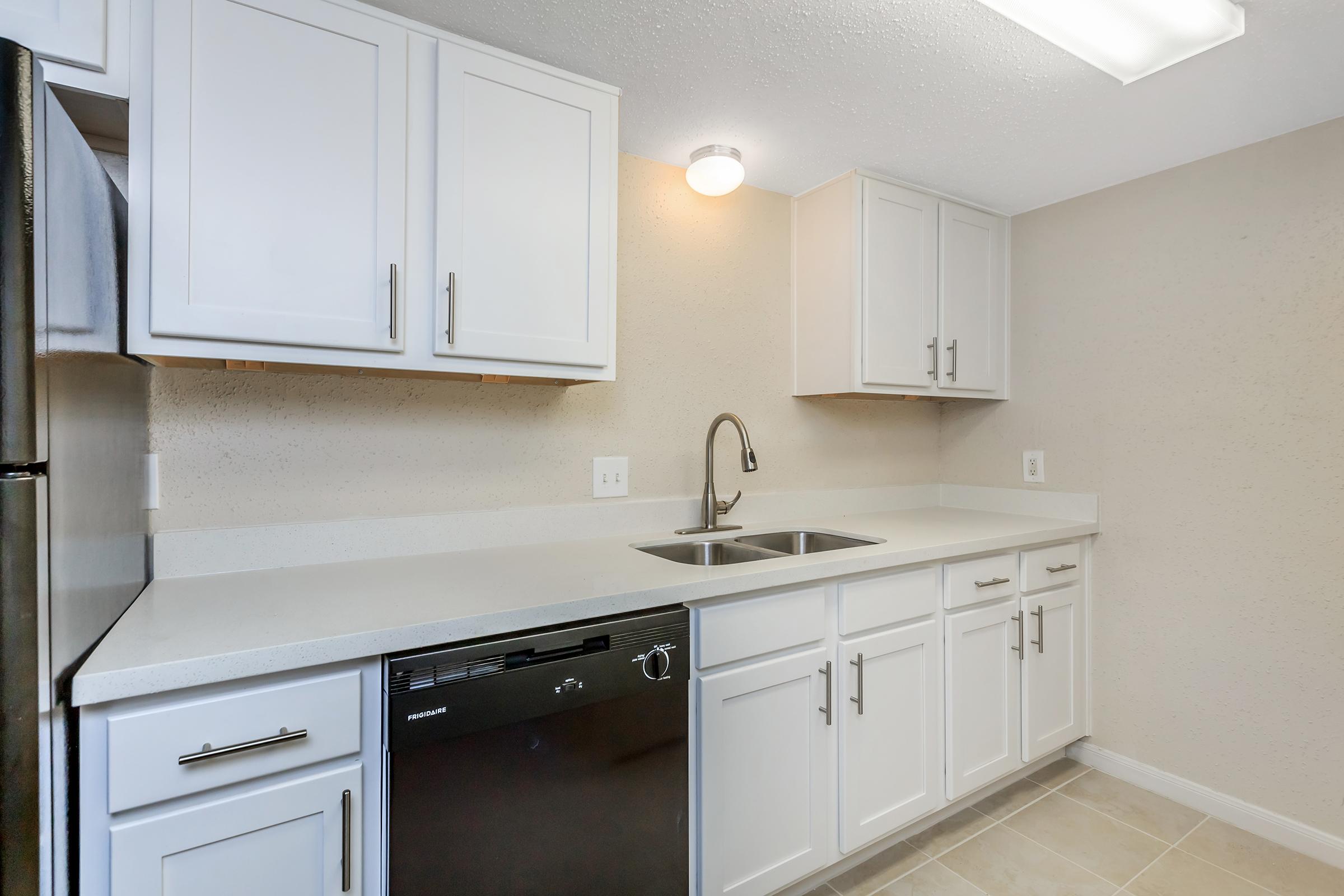 A modern kitchen featuring white cabinets, a stainless steel sink with a sleek faucet, a black dishwasher, and a refrigerator. The countertops are light-colored, and the walls are a neutral beige. The space is well-lit with overhead lighting, creating a clean and inviting atmosphere.