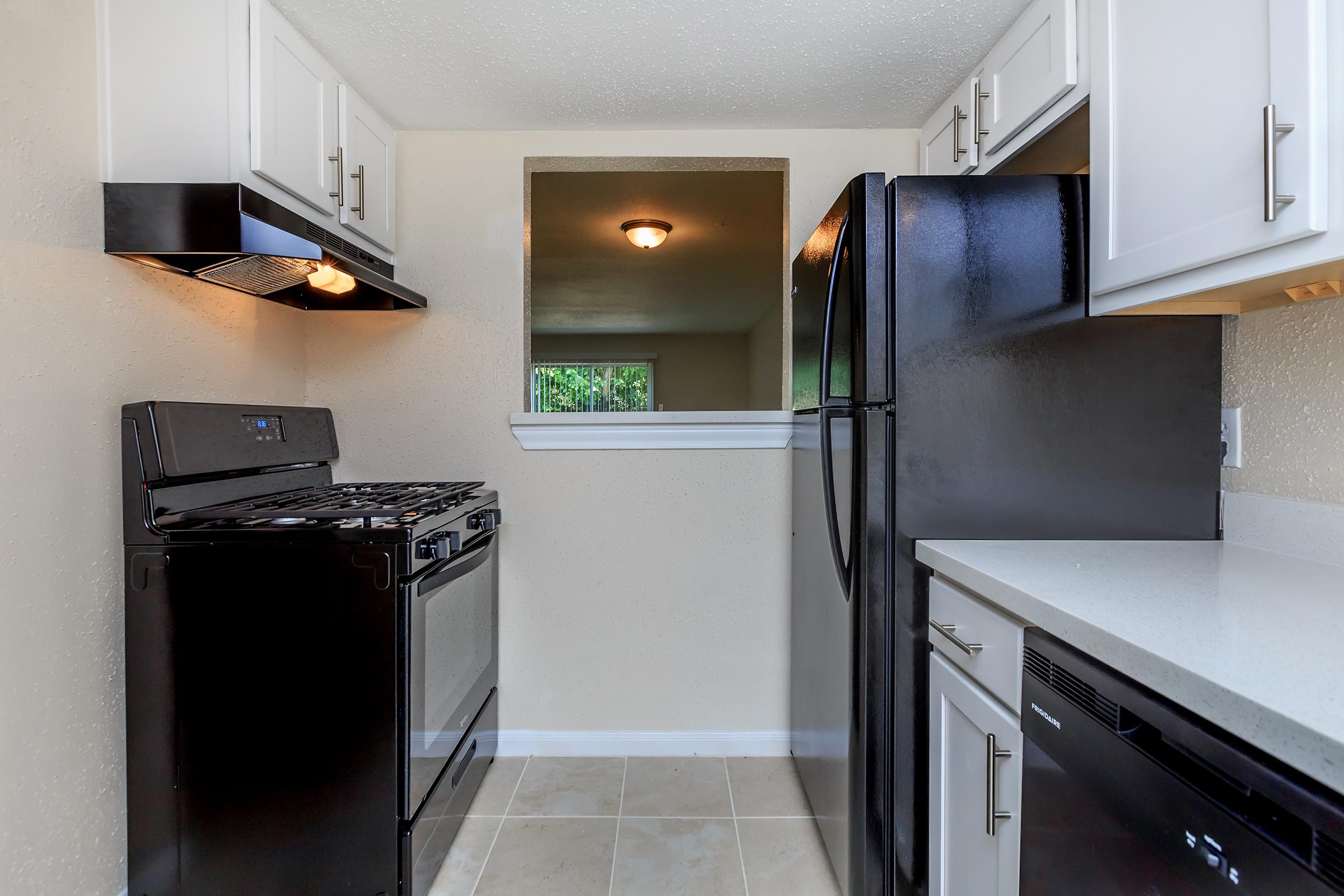 Modern kitchen featuring black appliances, including a gas stove, refrigerator, and dishwasher. White cabinets provide storage, and a window offers natural light. The countertop is light-colored, with a tile floor completing the clean, contemporary look. The space is compact yet functional, ideal for cooking and meal preparation.