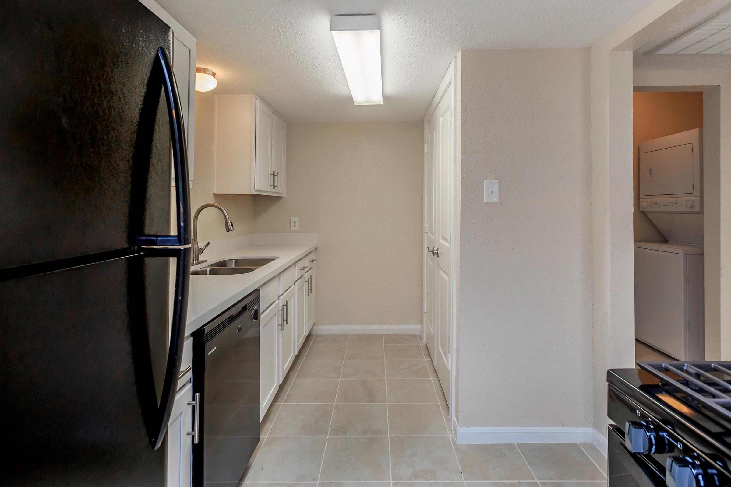 A modern kitchen featuring a black refrigerator and dishwasher, light-colored cabinets, and a sink. The space has beige walls and tiled flooring, with ample natural light from a ceiling fixture. A laundry area is visible through an open door at the back.