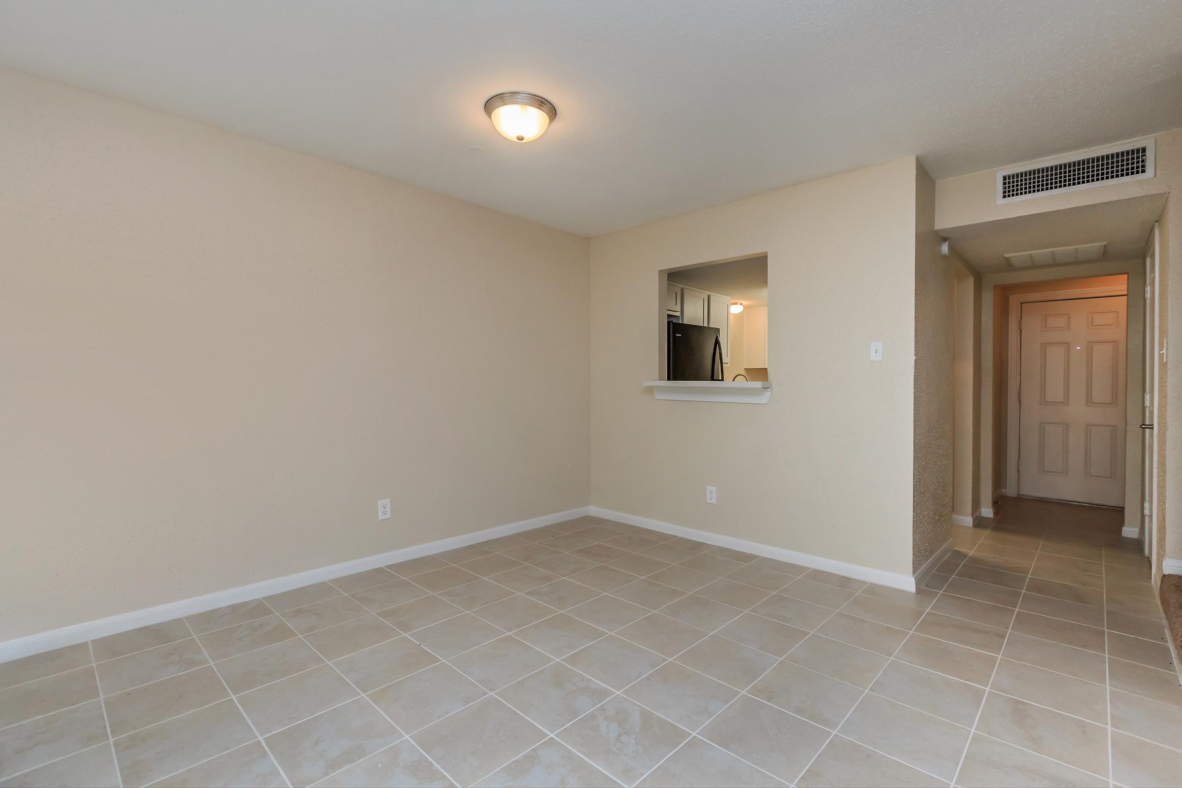 A spacious, empty room with beige walls and tiled flooring. There is a ceiling light fixture and an opening leading to a kitchen area in the back. A hallway is visible on the right, showing a door and additional space beyond. The overall ambiance is clean and unfurnished, ready for personalization.