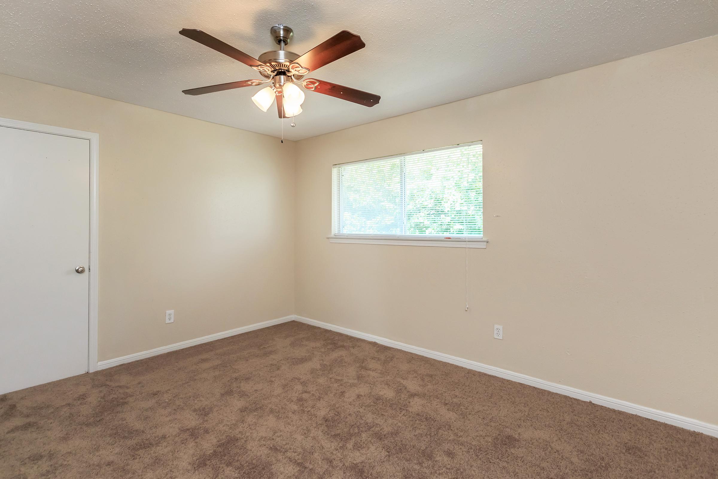 Empty room with light beige walls and a light brown carpet. A ceiling fan with three lights hangs from the ceiling, and there is a white door on one side. A window with blinds allows natural light to enter, illuminating the space. The overall atmosphere is bright and uncluttered.