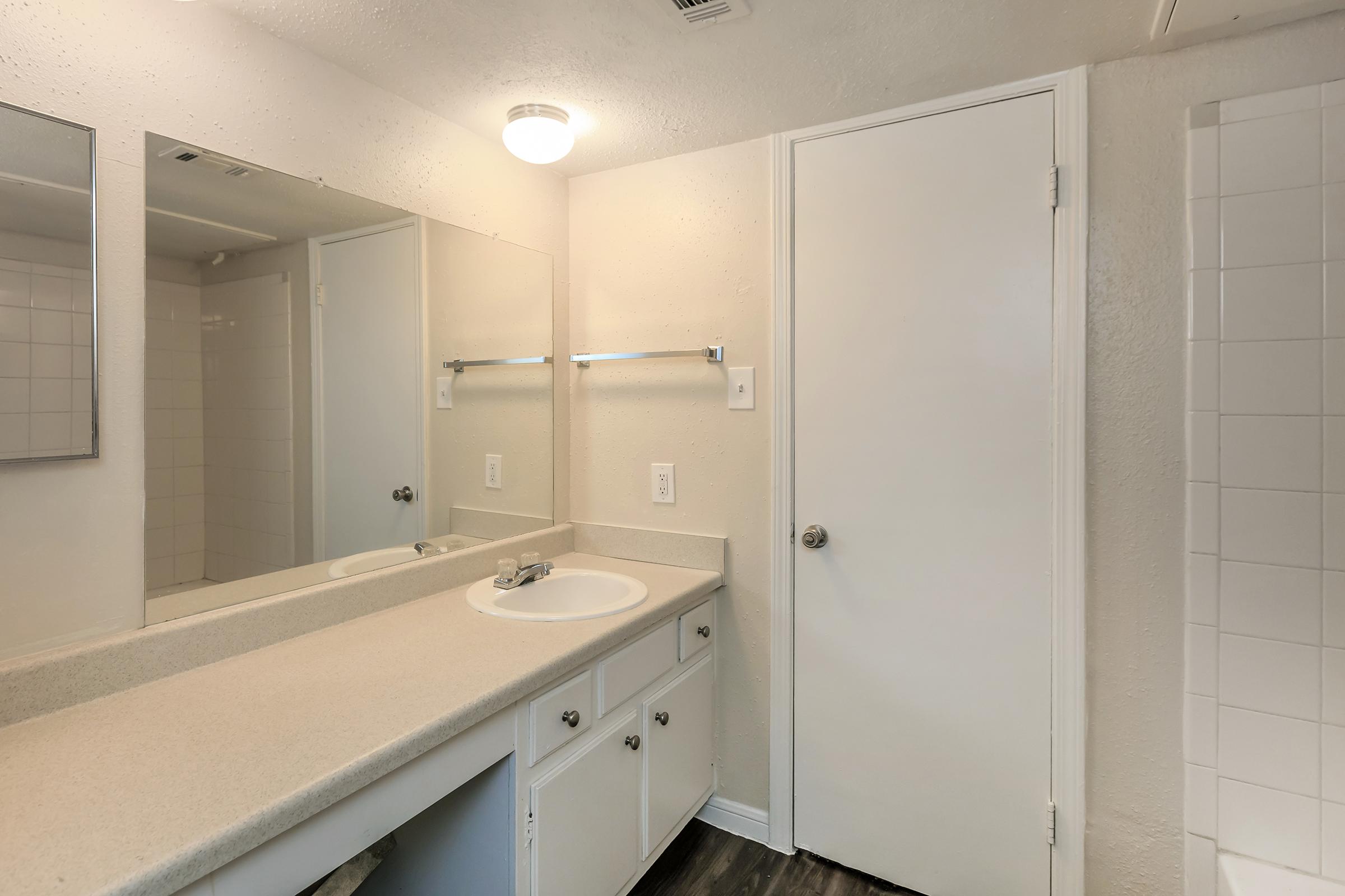 A bathroom featuring a light-colored countertop with a sink, a large mirror above, and a white cabinet below. There is a light fixture on the ceiling, a white door, and a tiled shower area visible in the background. The walls are painted in a light shade, creating a bright and clean atmosphere.