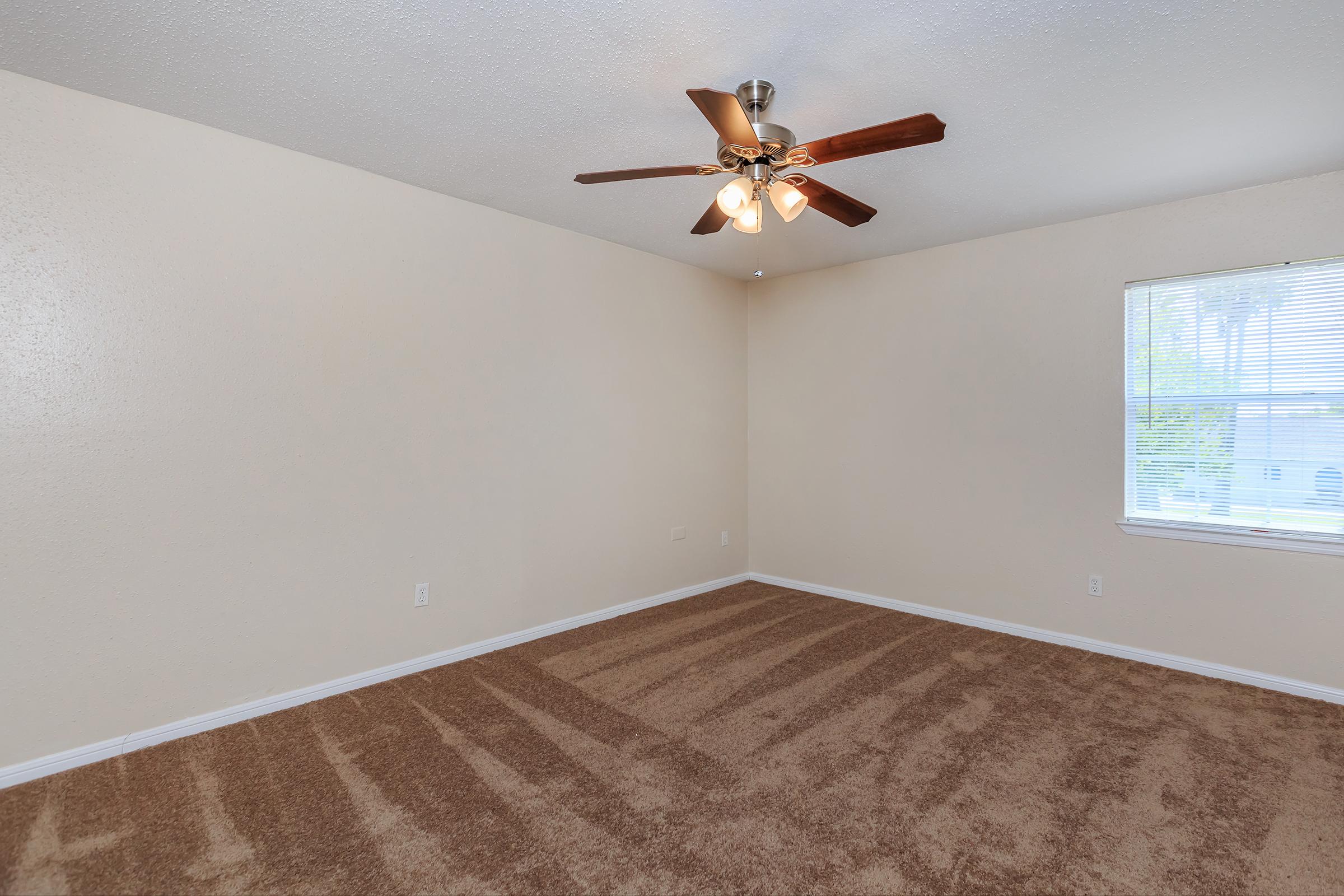 An empty room with light brown carpet, a ceiling fan with four light bulbs, and a window allowing natural light. The walls are painted in a neutral beige color, creating a spacious and clean atmosphere.