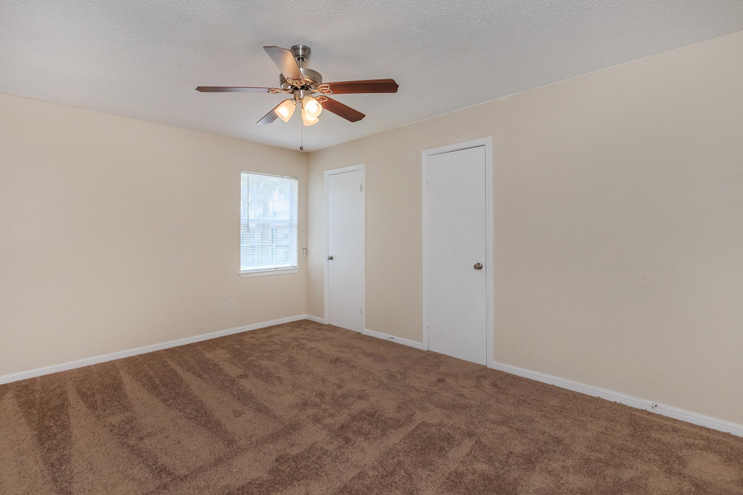 A cozy room featuring light beige walls and brown carpet. A ceiling fan with four light bulbs is installed, and there is a window allowing natural light. Two white doors are visible on the right, suggesting closet space. The overall atmosphere is warm and inviting.