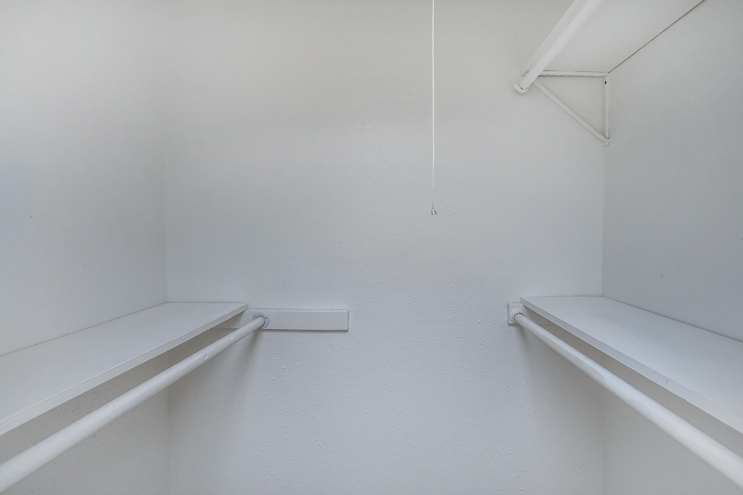 Empty white closet with two horizontal clothing rods and shelves on either side. The walls and ceiling are plain, emphasizing the minimalistic design. A single light pull chain hangs from the ceiling, adding to the overall simplicity of the space.