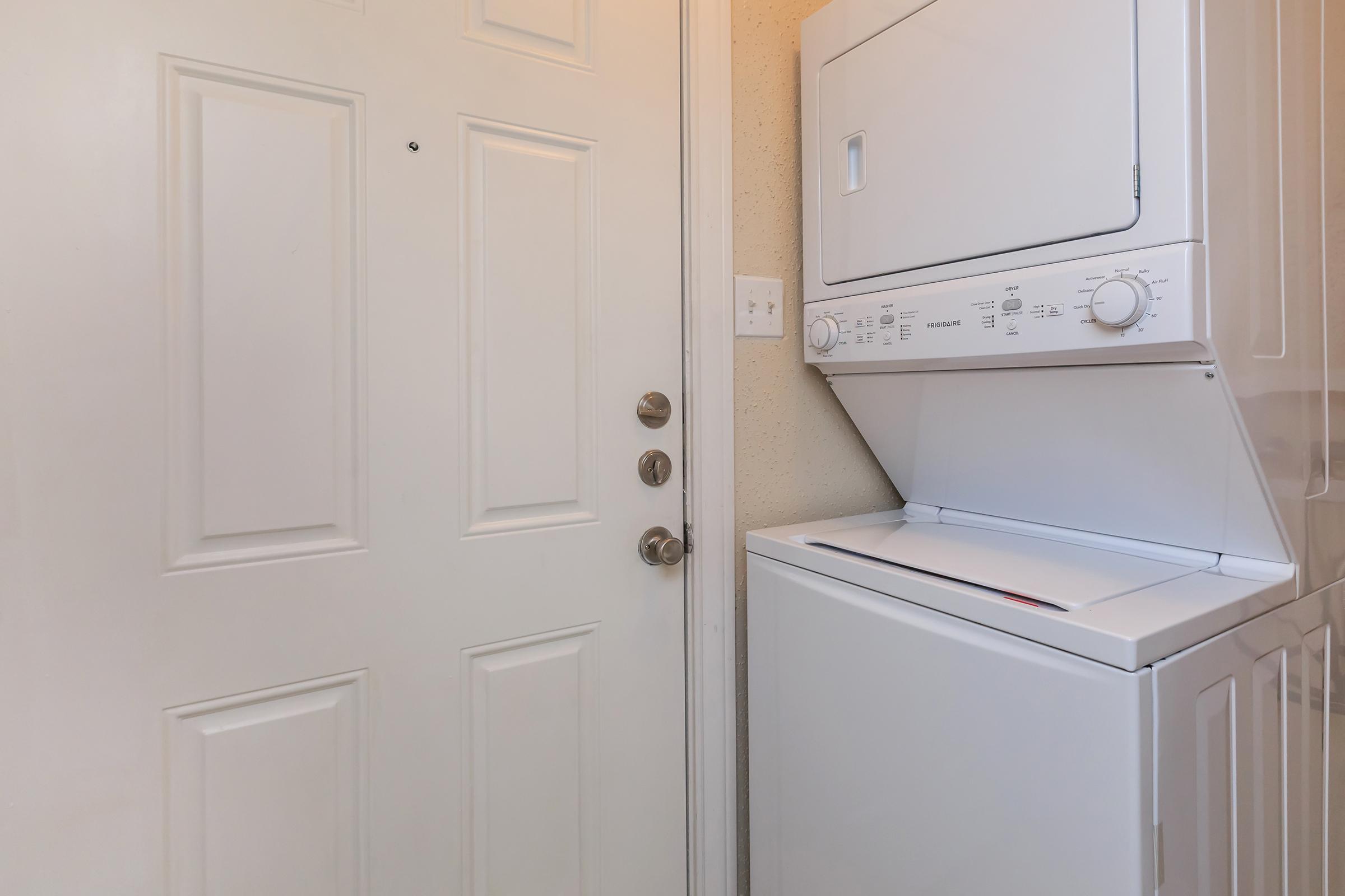 A stacked washer and dryer unit is positioned next to a closed door with a knob and a peephole. The laundry appliances are white, and the door is a light beige color with a simple design. The space appears tidy and functional, suitable for a laundry area in a home.