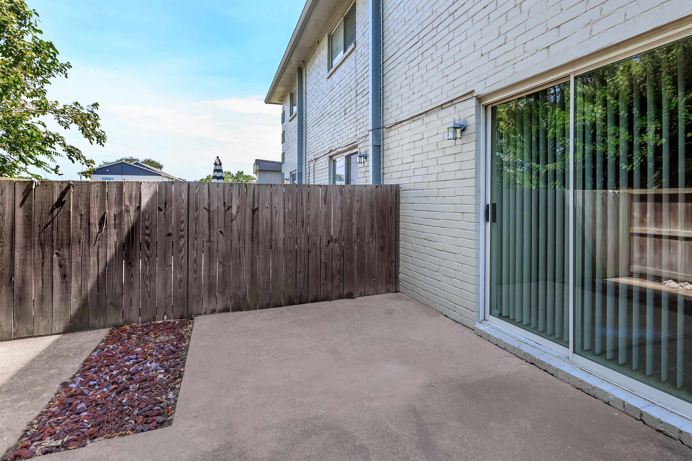 A small outdoor patio area featuring a concrete floor, a wooden fence to one side, and sliding glass doors leading into a building. The space is partially shaded with a few trees visible in the background and decorative gravel along the edge.