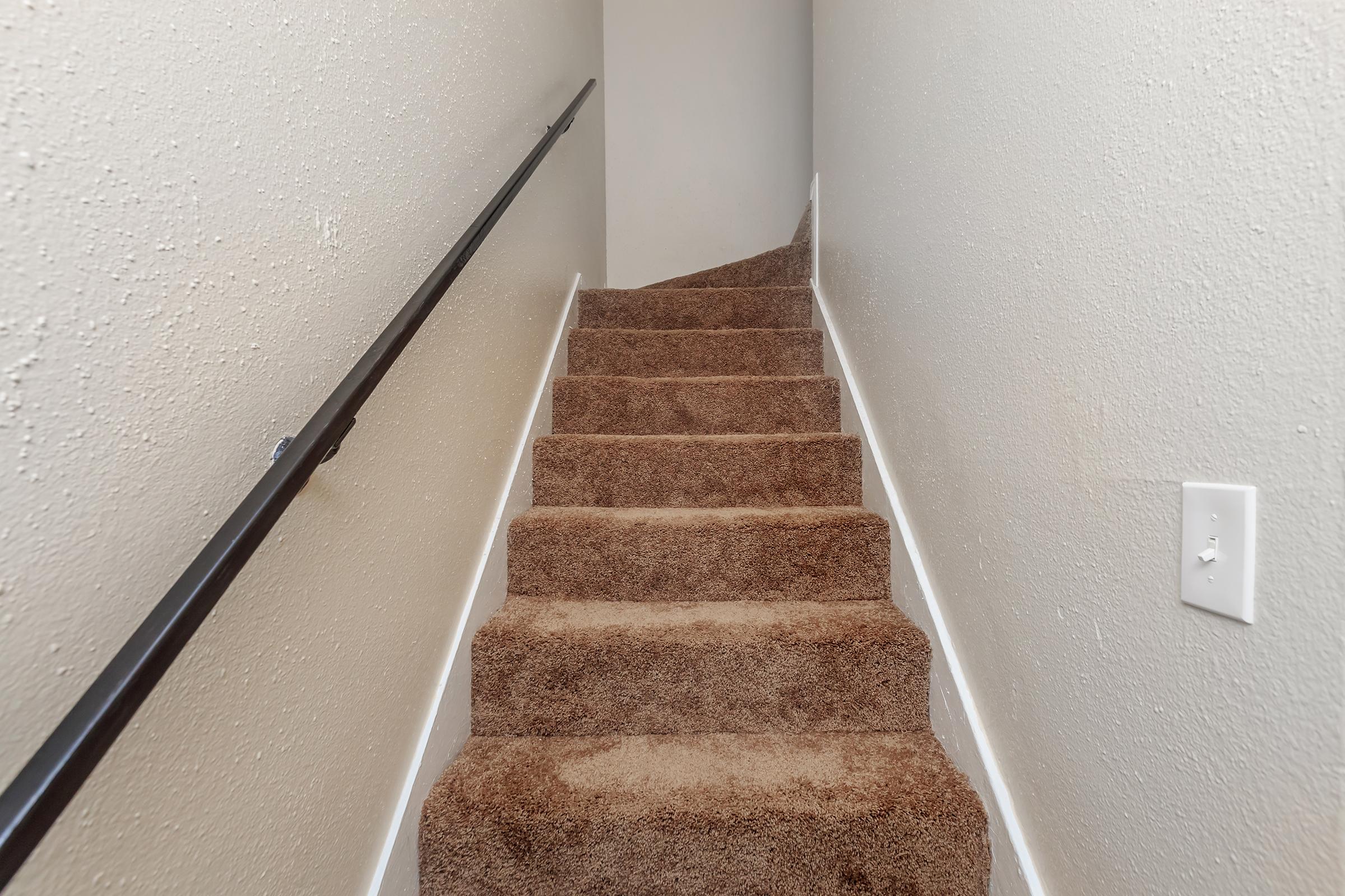 A narrow carpeted staircase leading upwards, with a simple handrail on one side and plain light-colored walls. The steps are covered in light brown carpeting, and there is a slight curve at the top of the staircase. The overall atmosphere is clean and inviting.