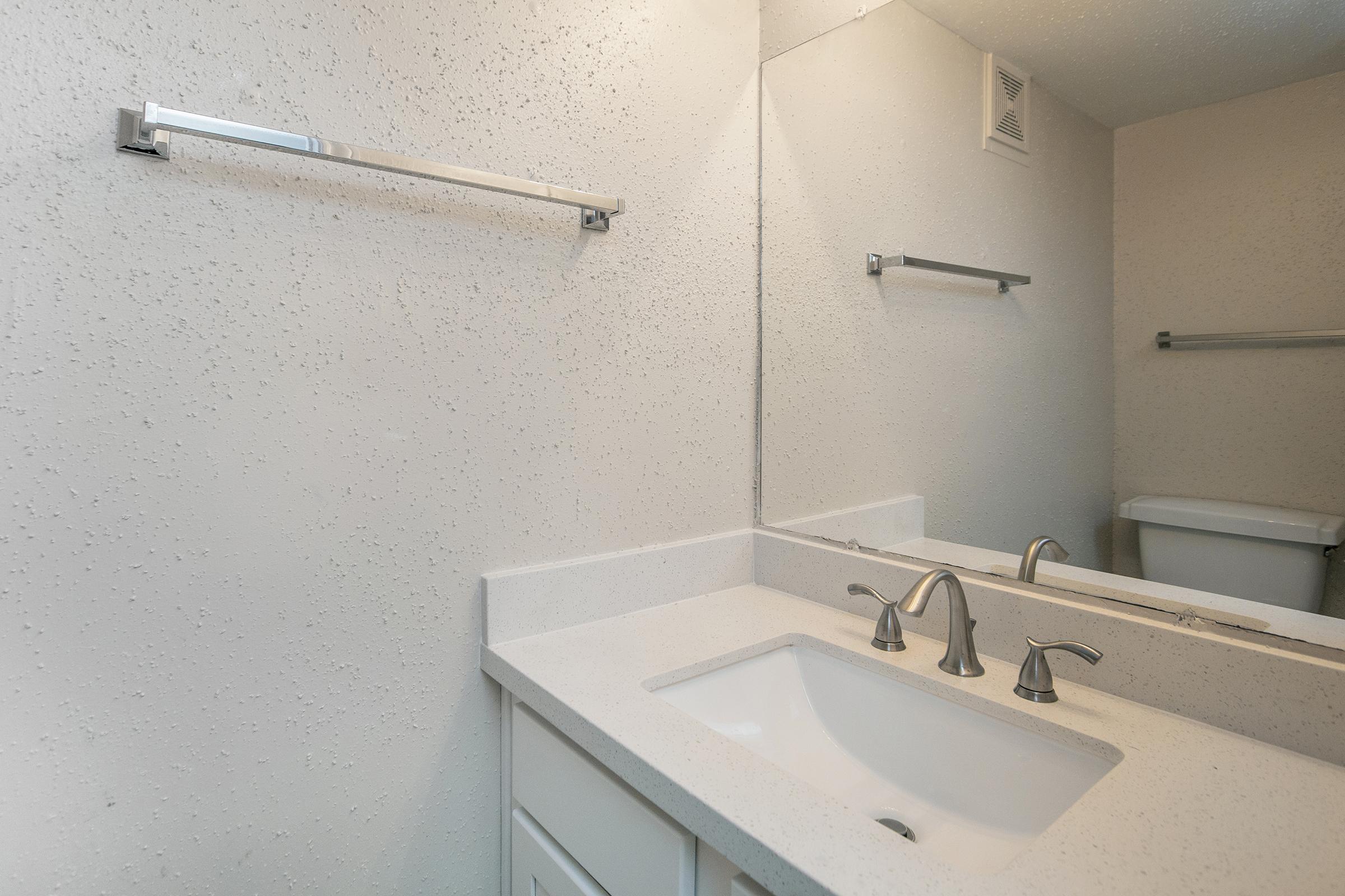 A clean bathroom interior featuring a white countertop with an under-mount sink and two chrome-finished faucets. A large mirror above the sink reflects the wall and an adjacent towel bar. The walls have a textured finish, and a toilet is visible in the background, partially obscured.