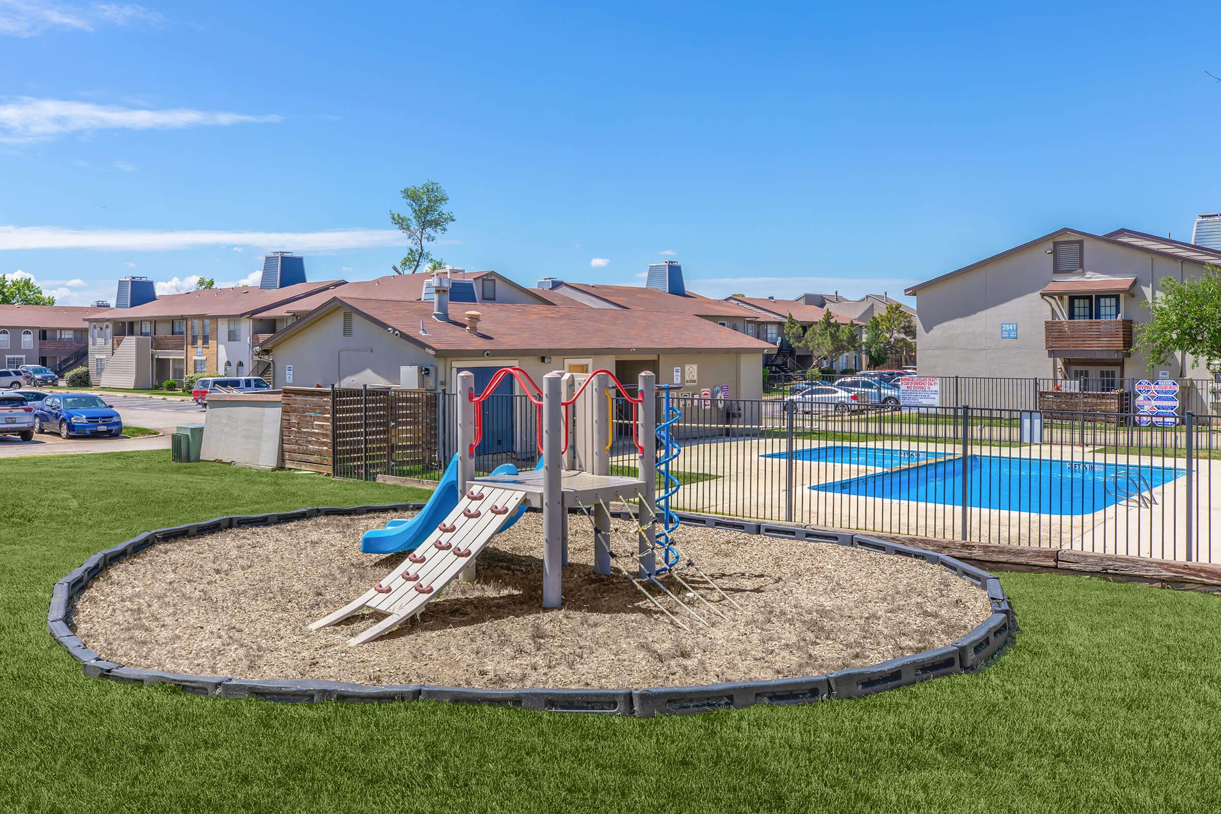 A playground featuring a slide and climbing structure, surrounded by mulch and a fence. In the background, there is a swimming pool and multiple apartment buildings under a clear blue sky.