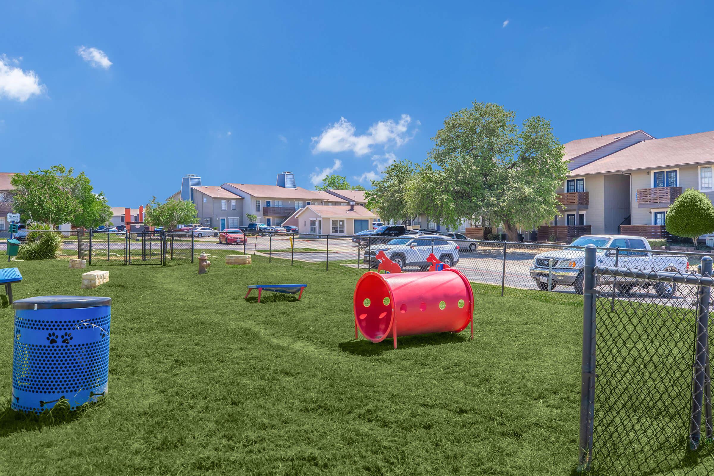 A bright outdoor dog park featuring green grass, a red tunnel, and various obstacles for dogs. Small benches and a blue trash can are visible, along with nearby residential buildings and parked cars under a clear blue sky.