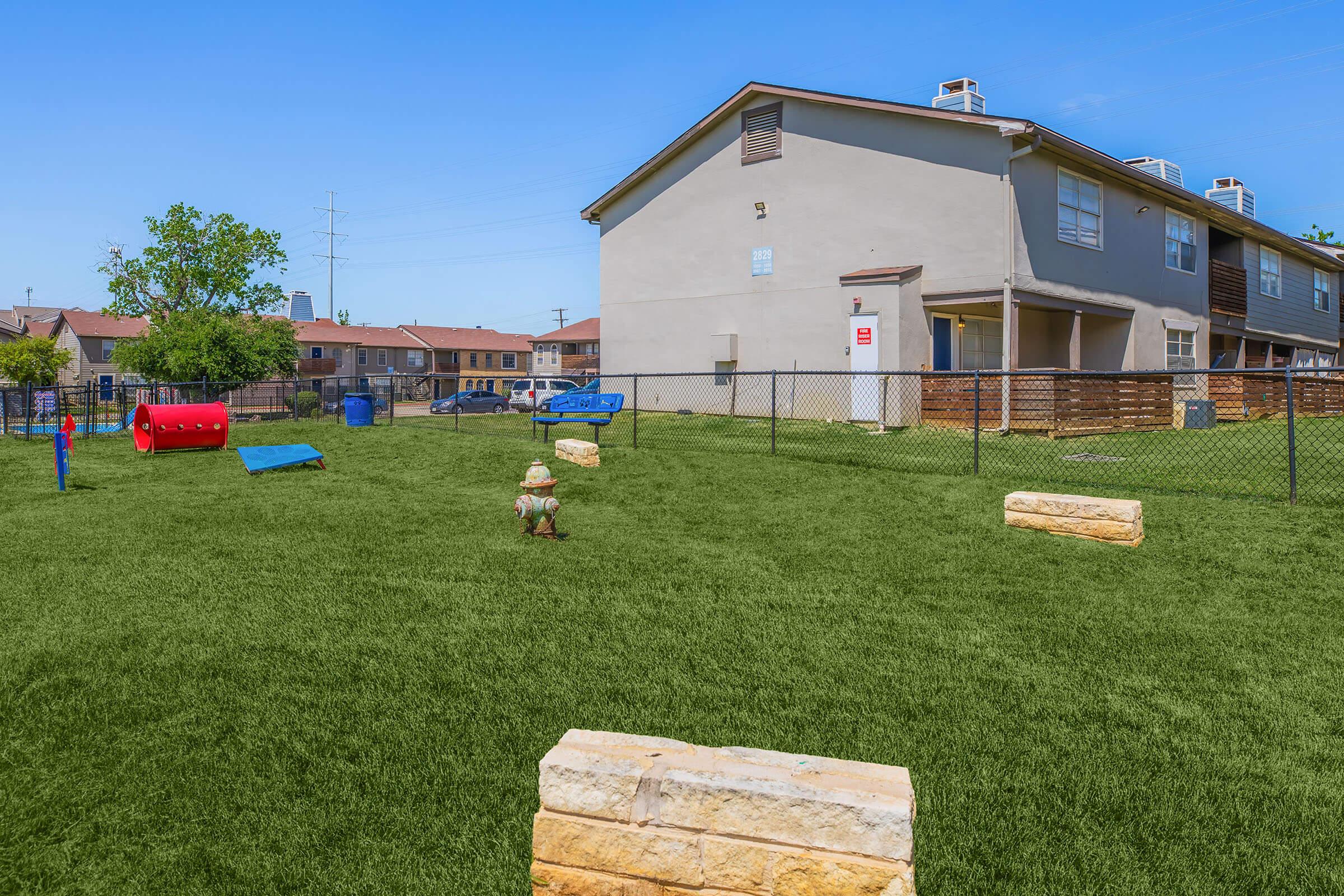 A vibrant playground area with artificial turf, featuring various play structures like slides and climbing frames. In the background, a residential building is visible, along with a fenced perimeter and green trees, creating a cheerful environment for children to play and explore.
