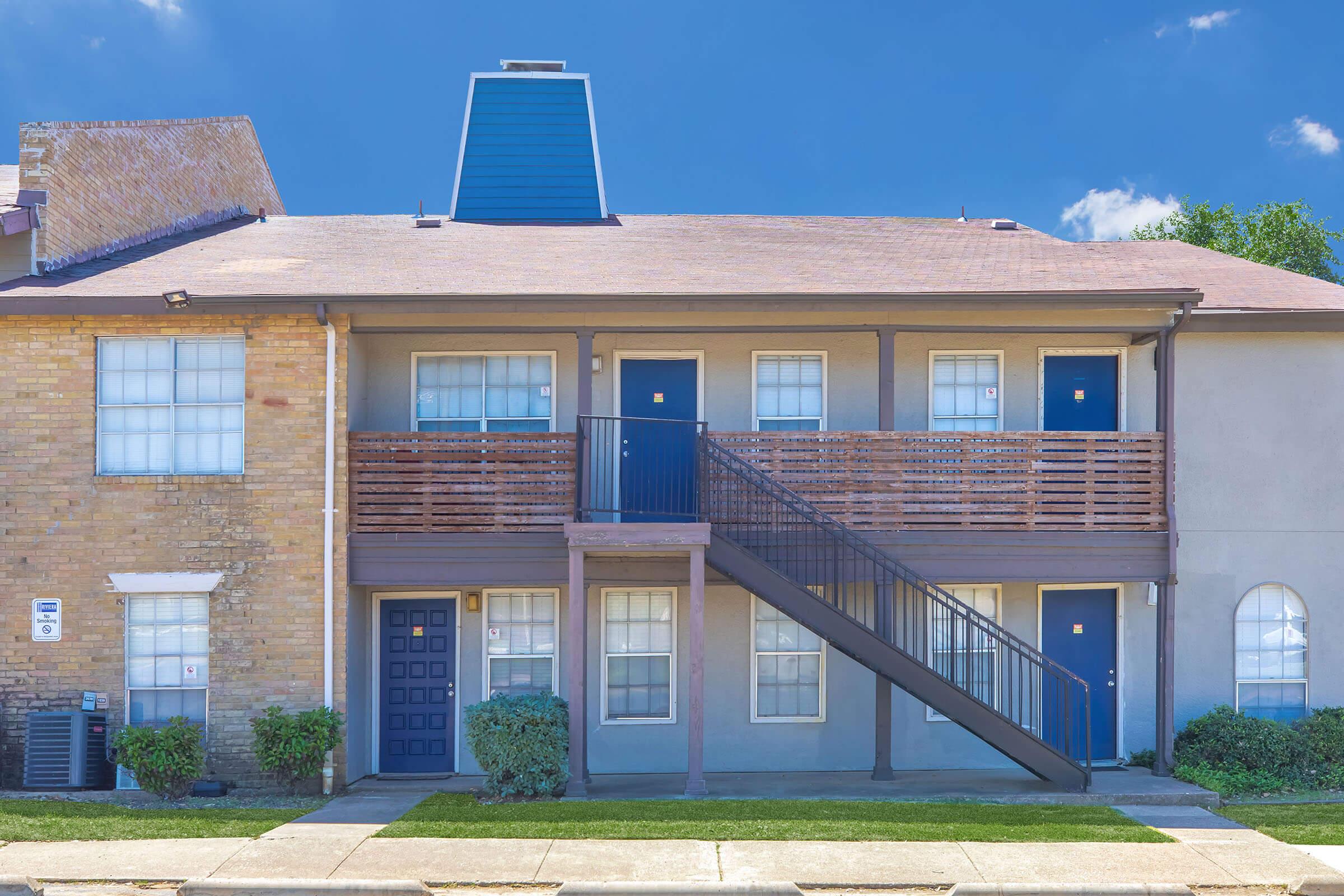 A two-story apartment building with a symmetrical facade. The left side features a brick exterior, while the right side is painted in a light color. Each unit has a blue door, and a staircase leads to the second floor. Green shrubs line the walkway, and the sky is clear with few clouds.
