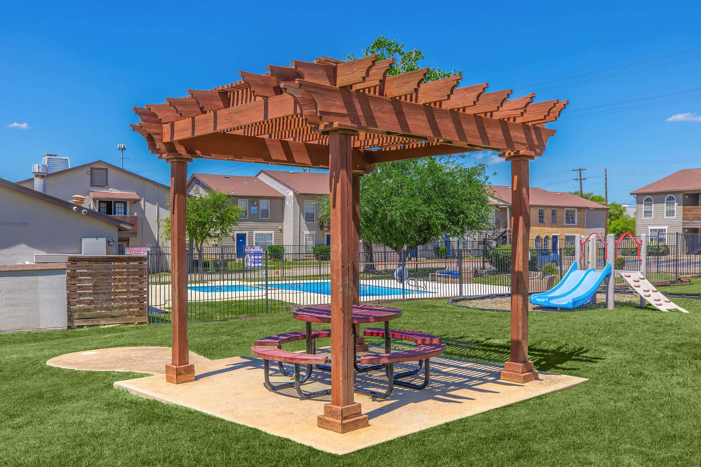 A wooden pergola with a picnic table underneath, positioned on a grassy area. In the background, there is a fenced swimming pool and playground equipment, including slides. The sky is blue and clear, with a few trees visible nearby.