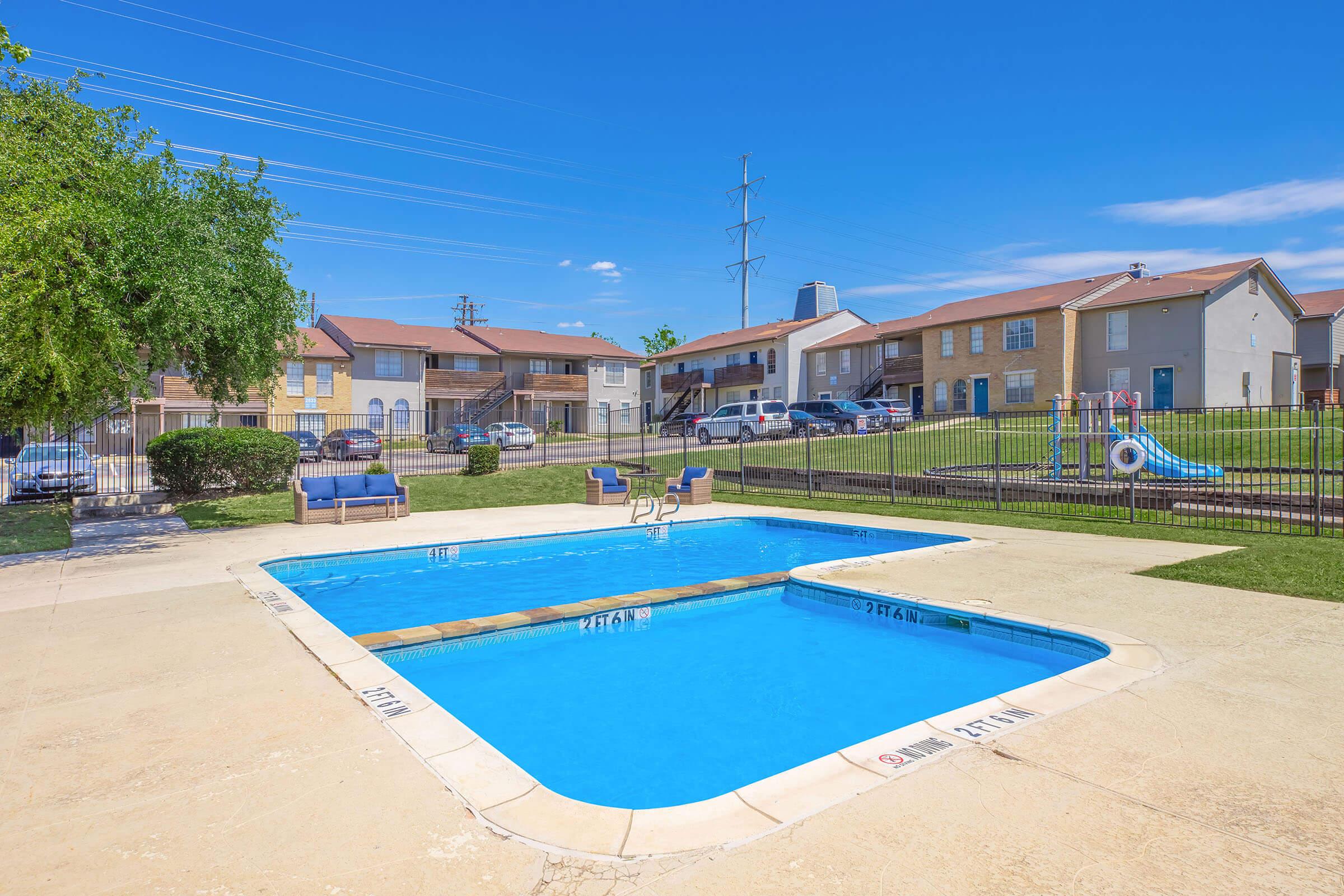 A view of a bright blue swimming pool surrounded by a concrete deck. In the background, there are two-story apartment buildings and a grassy area. There are a few lounge chairs near the pool, and a playground structure is visible to the side. Clear blue skies overhead complete the scene.