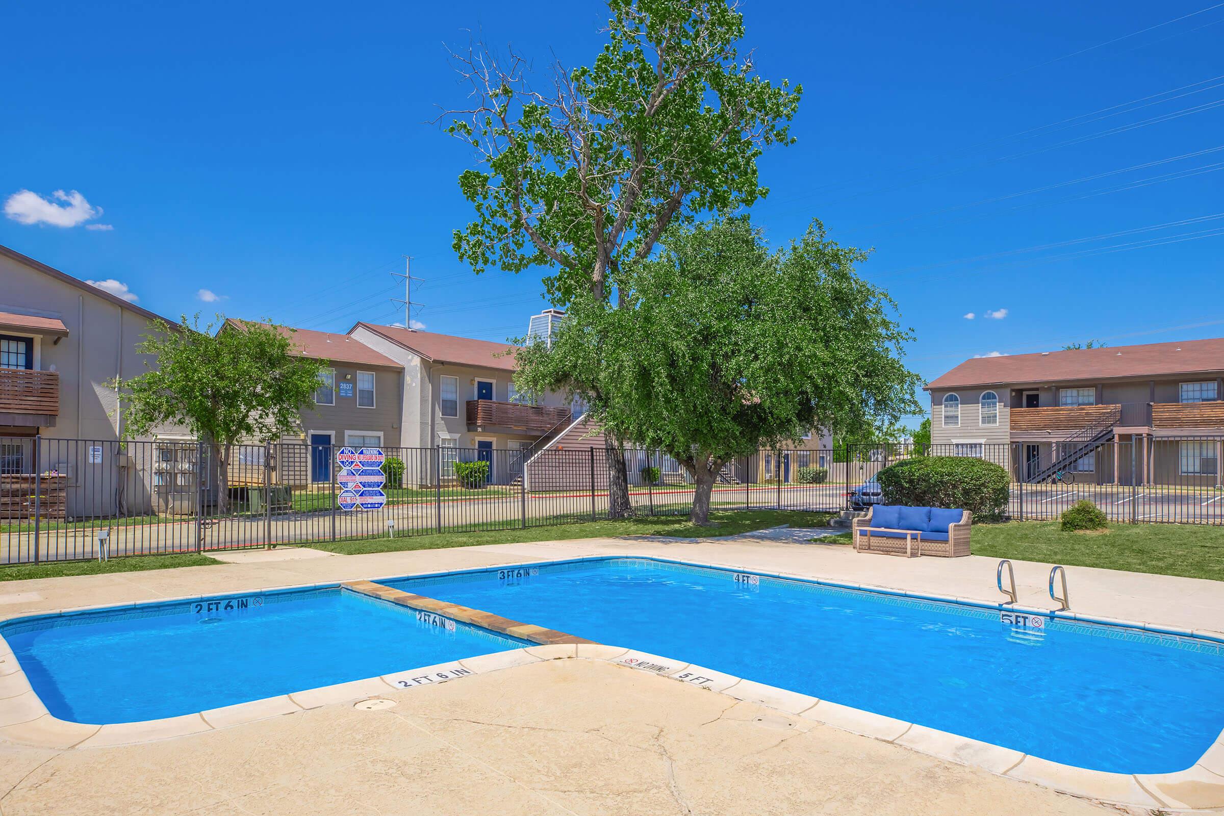 A sunny outdoor swimming pool area featuring two pools with clear blue water, surrounded by green grass and trees. In the background, there are residential buildings with balconies. A lounge chair and signs are visible near the pool area. Clear blue sky adds to the inviting atmosphere.