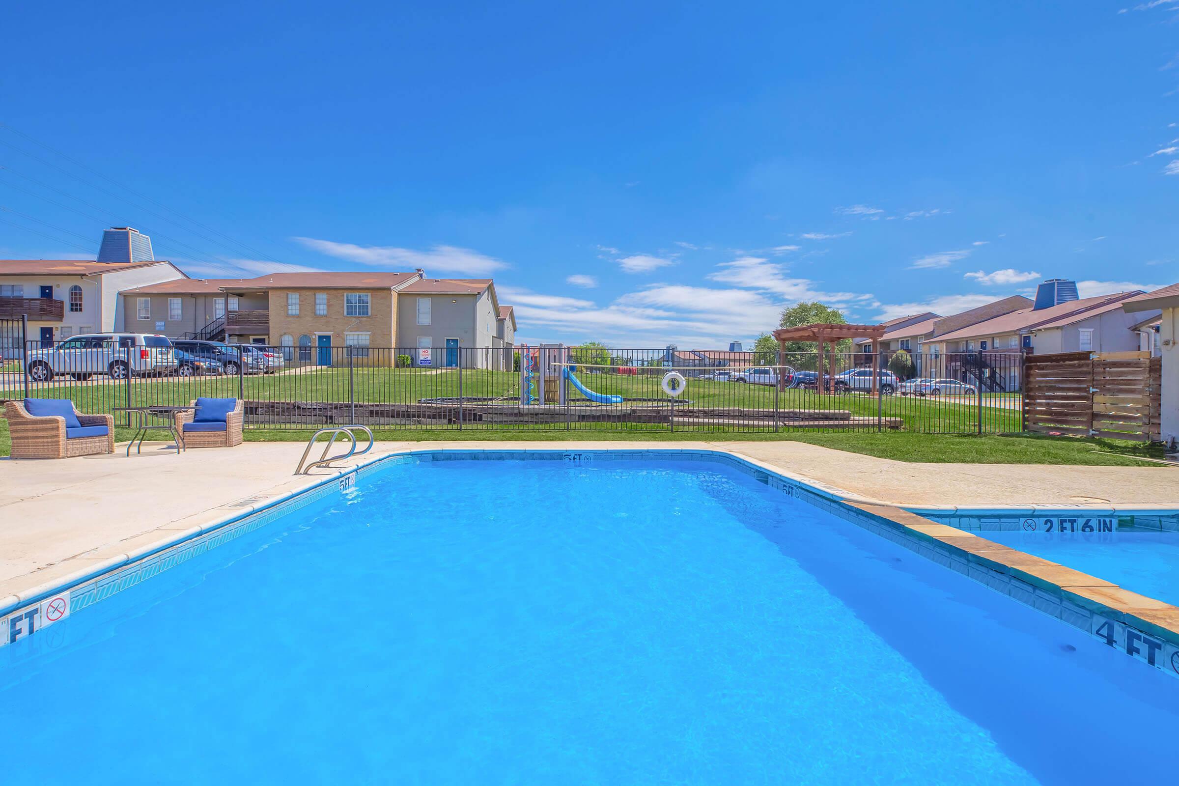 A clear blue swimming pool with lounge chairs on the edge, surrounded by a fence. In the background, there are several residential buildings and a grassy area with a playground slide. The sky above is bright blue with a few clouds, creating a sunny and inviting atmosphere.