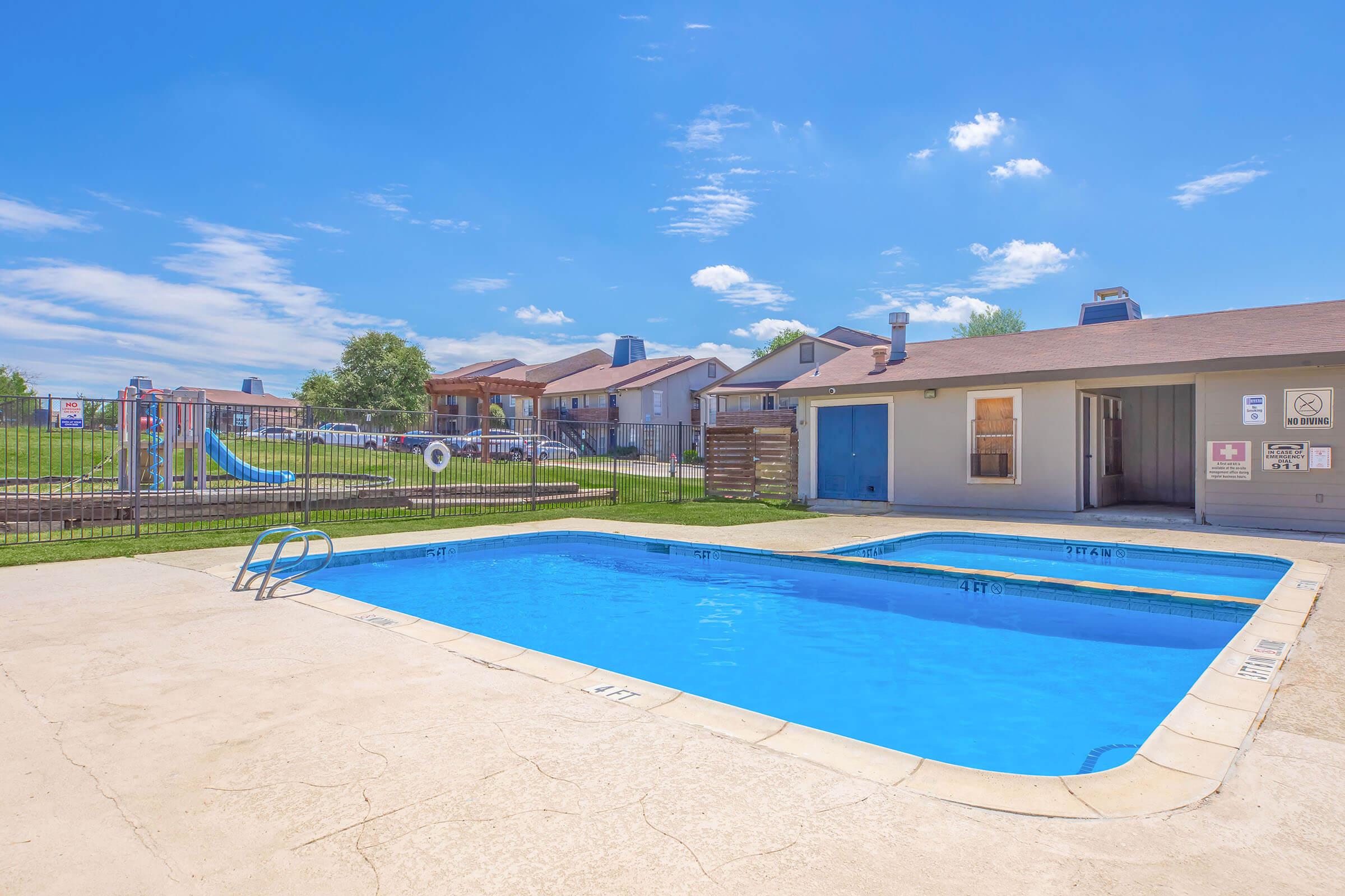 A clear blue swimming pool surrounded by a concrete deck, featuring a shallow and deep end. In the background, there are residential buildings and a playground with a slide. The sky is bright with a few fluffy clouds, creating a sunny atmosphere.