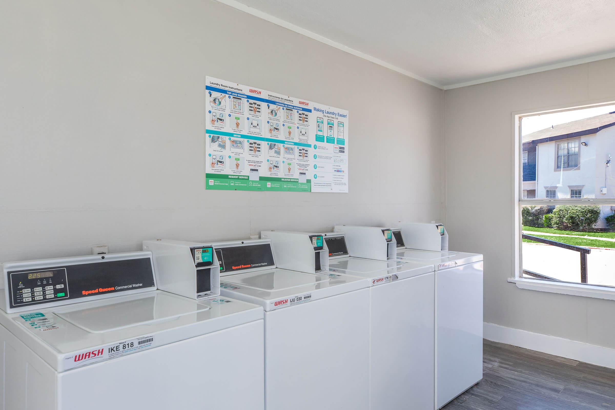 A clean and well-lit laundry room featuring four white washing machines lined up against a wall, a poster with laundry instructions and information displayed above them, and a window providing natural light with a view of the outside.