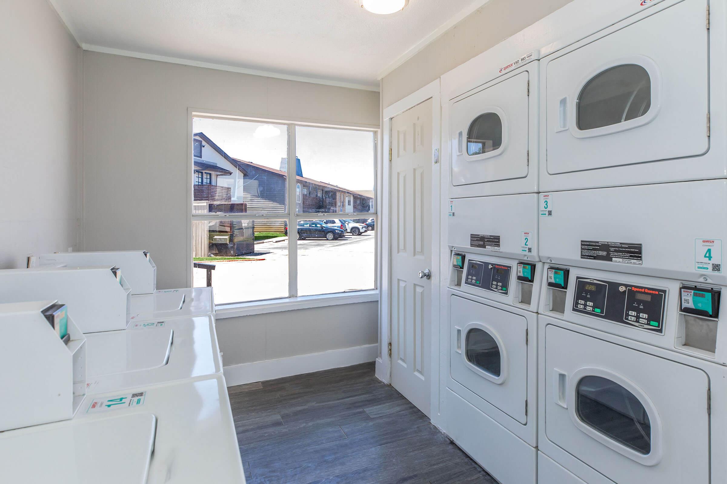 A bright laundry room featuring several white washing machines and dryers arranged along a wall. A large window allows natural light to enter, providing a view of the outside area. The flooring is dark, and the room has a clean, functional design.