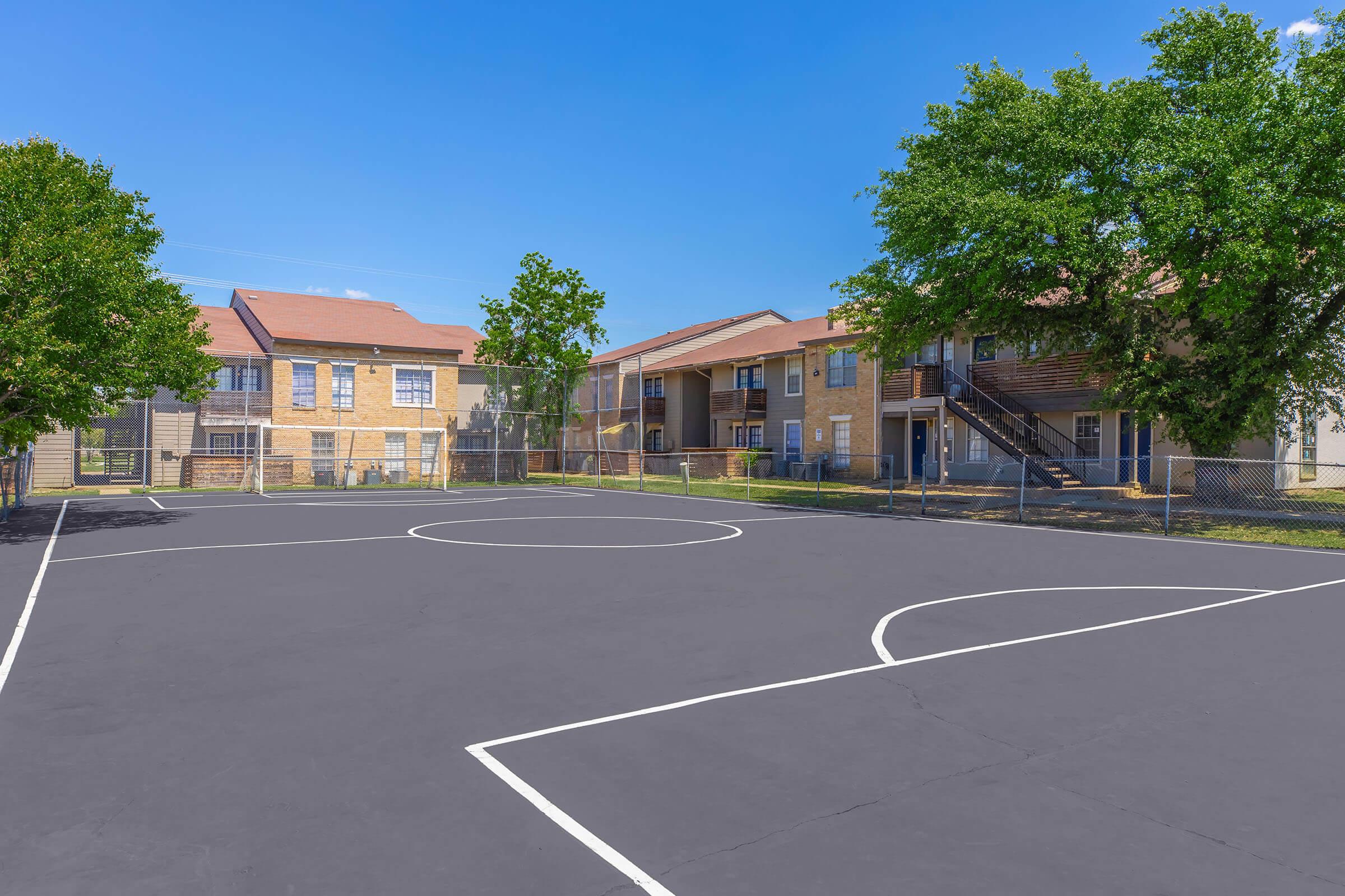 A basketball court with marked lines, surrounded by trees and two-story apartment buildings. The sky is clear and blue, creating a bright atmosphere. The court is fenced, and the area appears well-maintained, suggesting a neighborhood space for residents to enjoy outdoor activities.