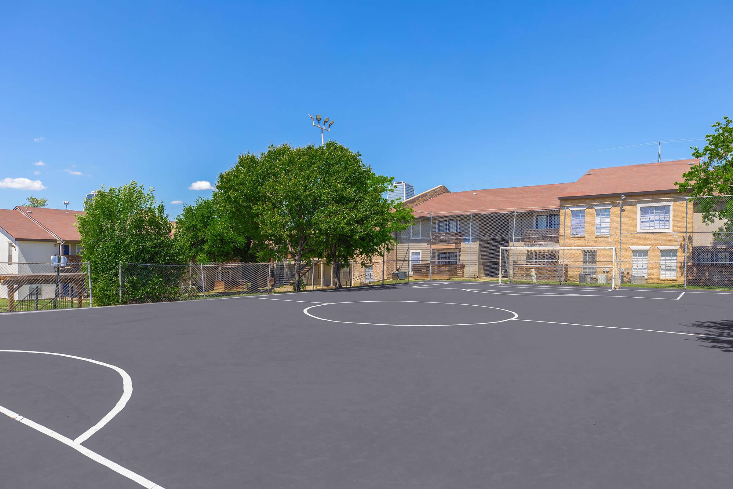 A sunny outdoor basketball court with a smooth black surface, featuring a half-court line and three-point arc. In the background, there are trees and a school building with a red roof, surrounded by a chain-link fence. The sky is clear and blue, creating a bright atmosphere.