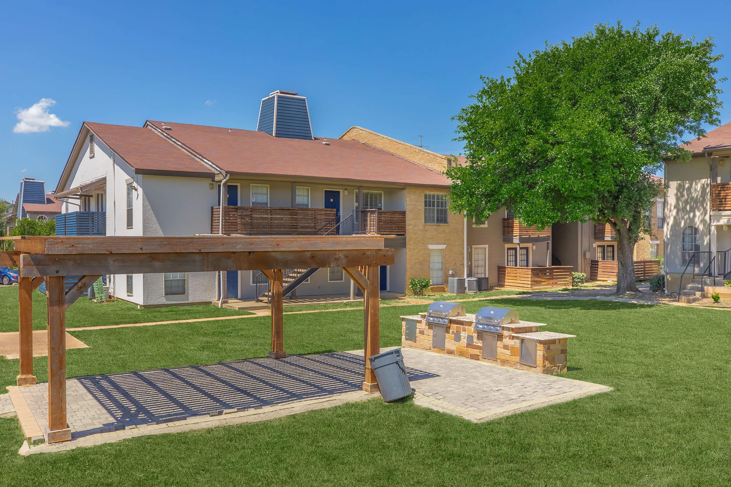 A well-maintained outdoor space featuring a wooden pergola, grill area with stone seating, and green grass. In the background, there are two multi-unit apartment buildings under a clear blue sky, with trees providing shade. Ideal setting for social gatherings or relaxation.