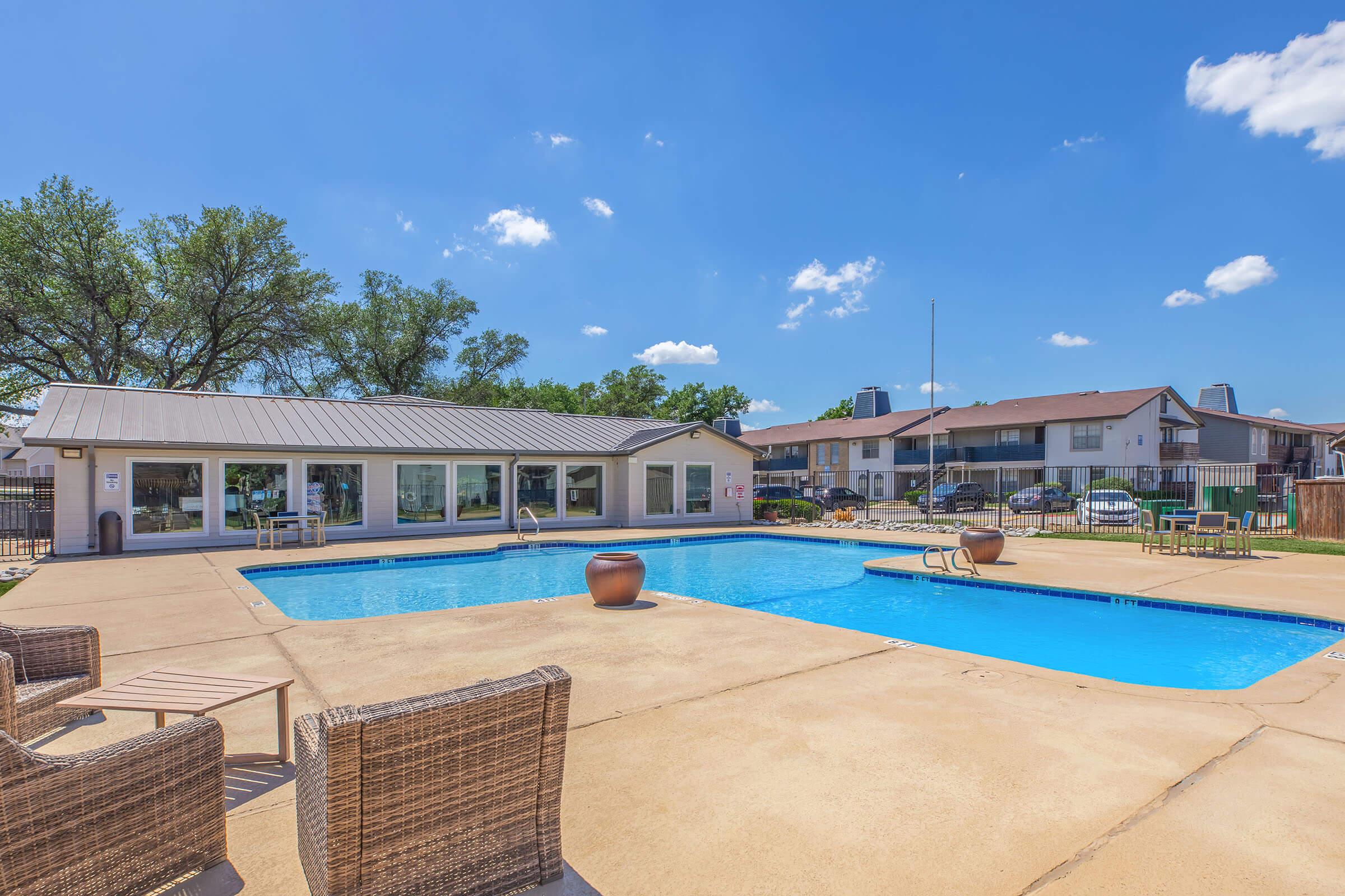 A sunny outdoor pool area featuring a rectangular swimming pool, lounge chairs, and a nearby clubhouse. Surrounding trees offer shade, with residential buildings visible in the background under a clear blue sky with a few clouds.