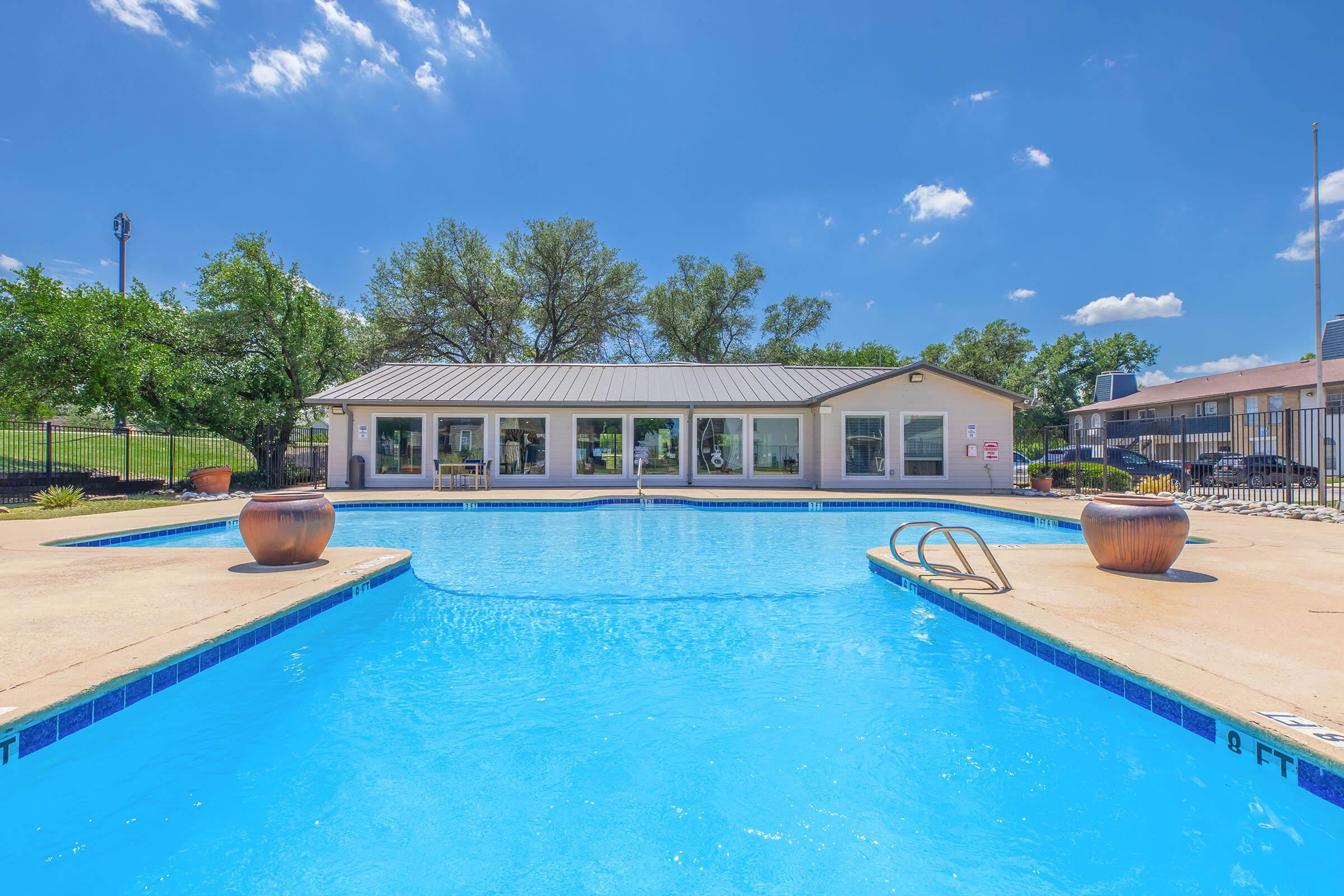 A sunny pool area featuring a clear blue swimming pool, surrounded by a deck and planters. In the background, there is a clubhouse with large windows. Lush green trees line the space, and a bright blue sky with a few fluffy clouds can be seen overhead.