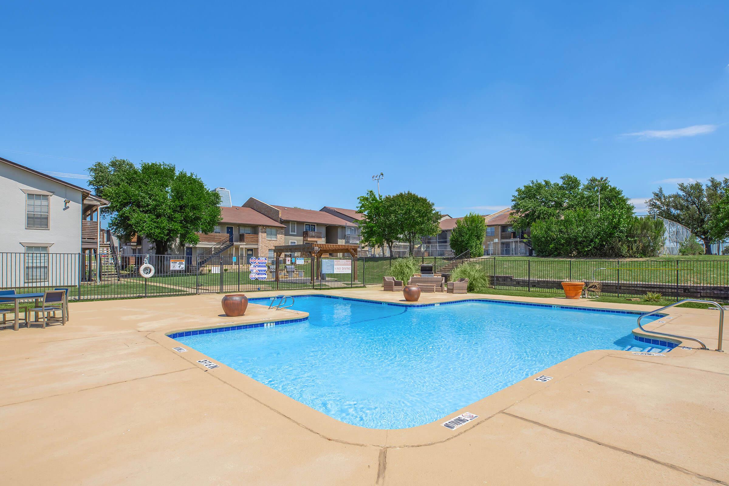 A clear blue swimming pool surrounded by a fenced area, with lounge chairs and potted plants. In the background, there are residential buildings with trees and grass. The sky is bright and sunny, creating a relaxing atmosphere.