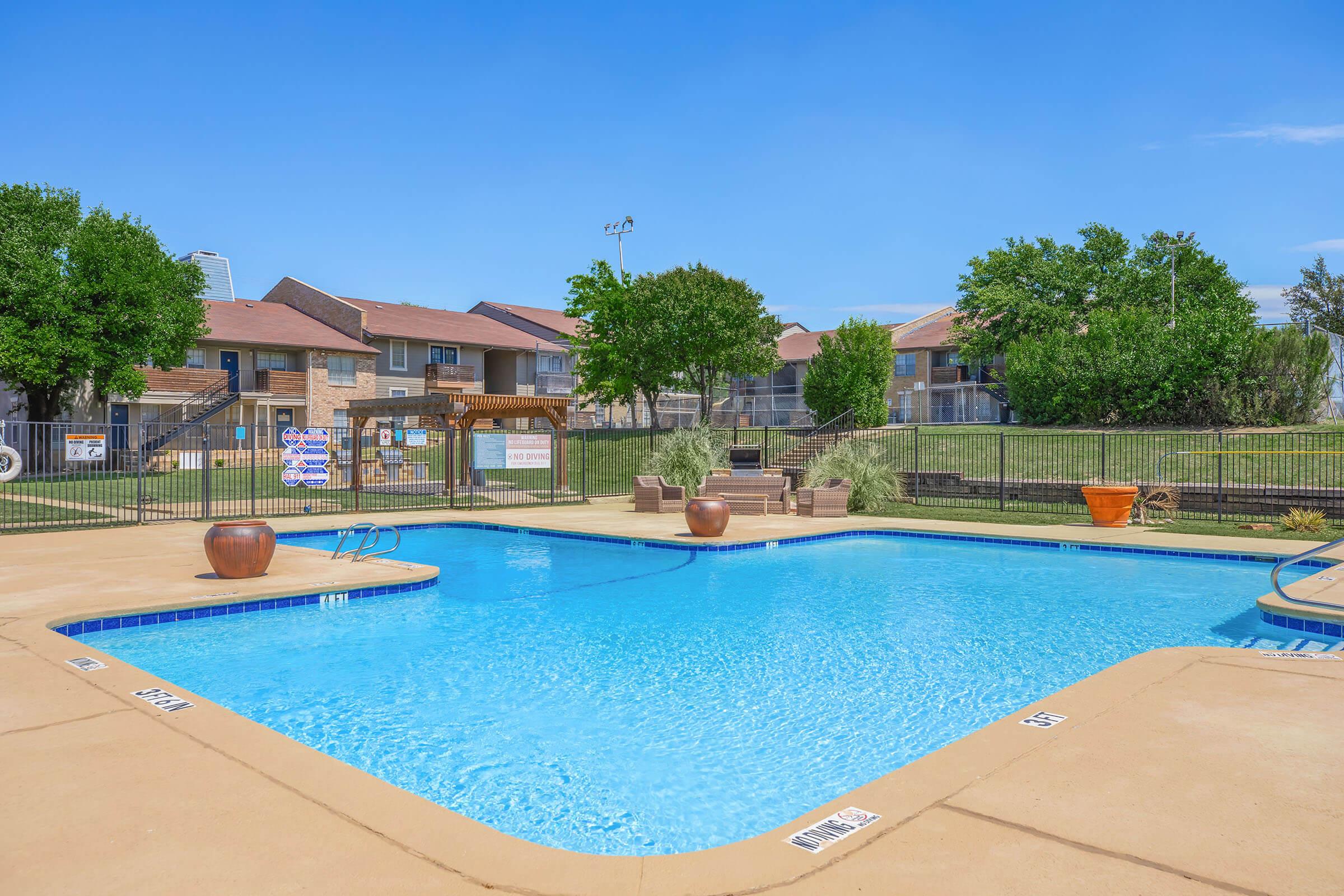 A clear blue swimming pool surrounded by a tan deck, with lounge chairs and potted plants. In the background, there are two-story apartment buildings and green trees under a bright blue sky. The pool area is fenced for privacy, and there is a pergola with seating nearby.