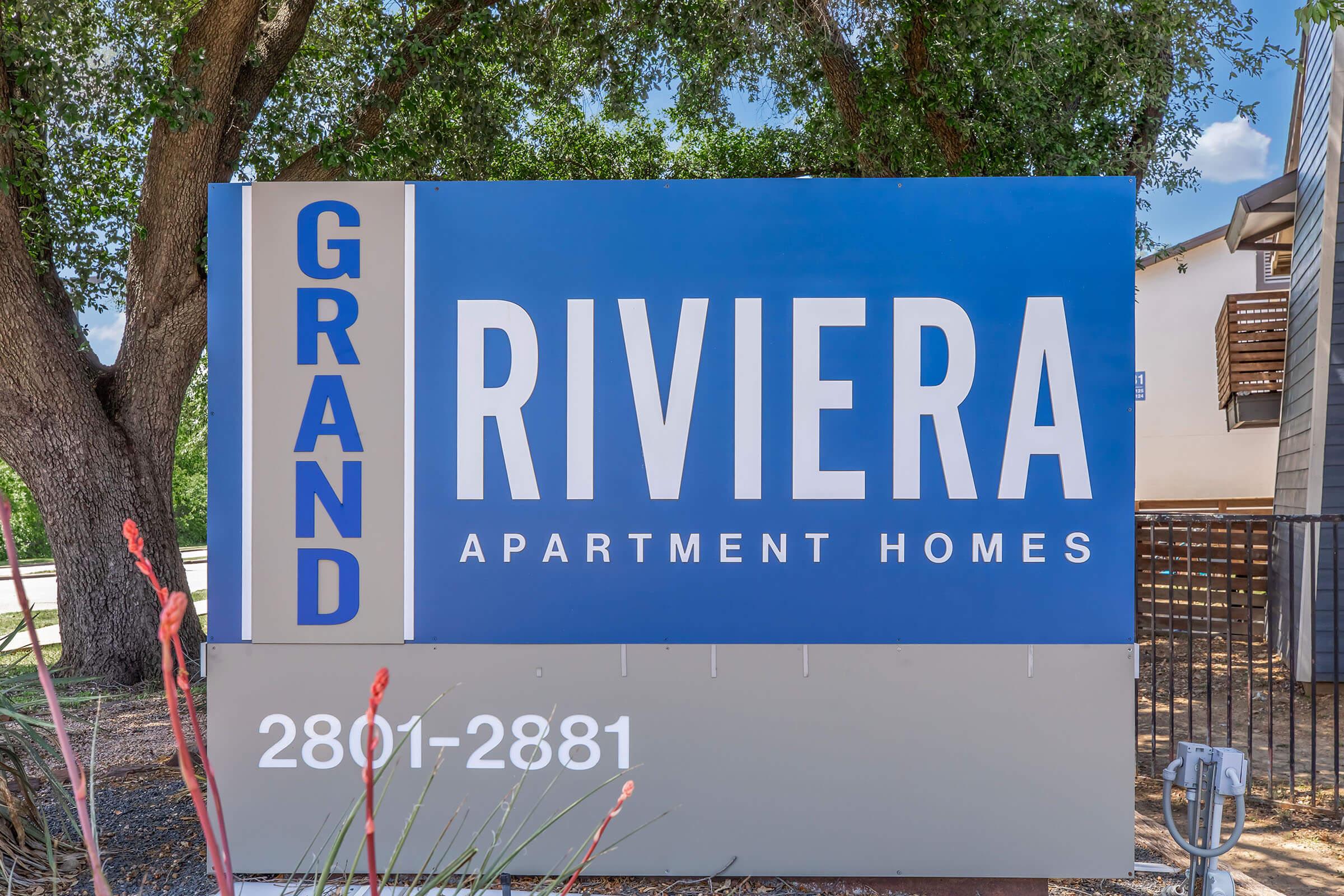 Sign for Grand Riviera Apartment Homes, displaying the name prominently in blue and white text. The address is 2801-2881, surrounded by greenery and a landscaped area. The sign is located in an outdoor setting with trees and plants nearby.