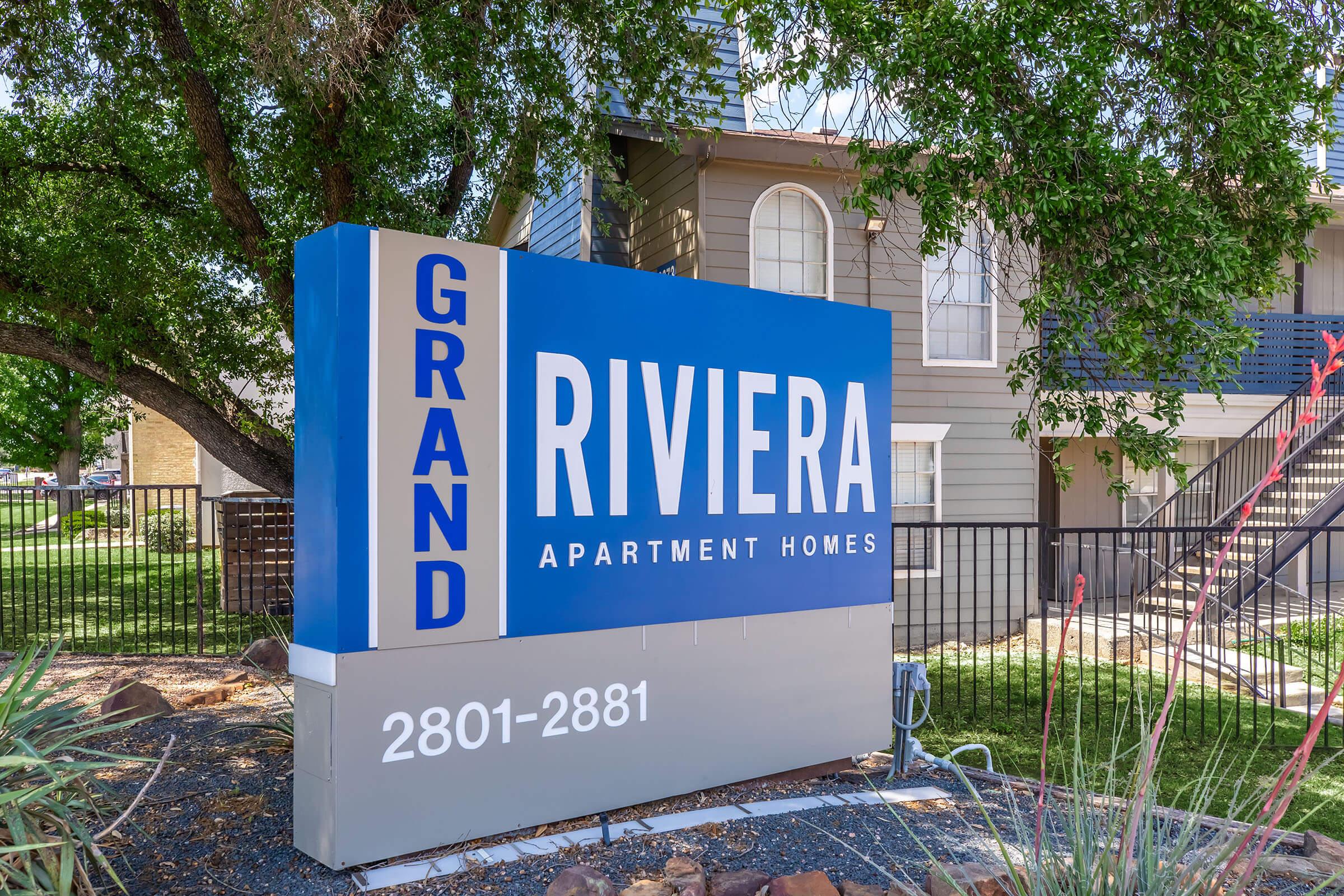Sign for Grand Riviera Apartment Homes, featuring a blue background with white text. The sign displays the name "Grand Riviera" prominently, along with the address range "2801-2881" below. Surrounding the sign are green trees and landscaping, indicating a residential area.