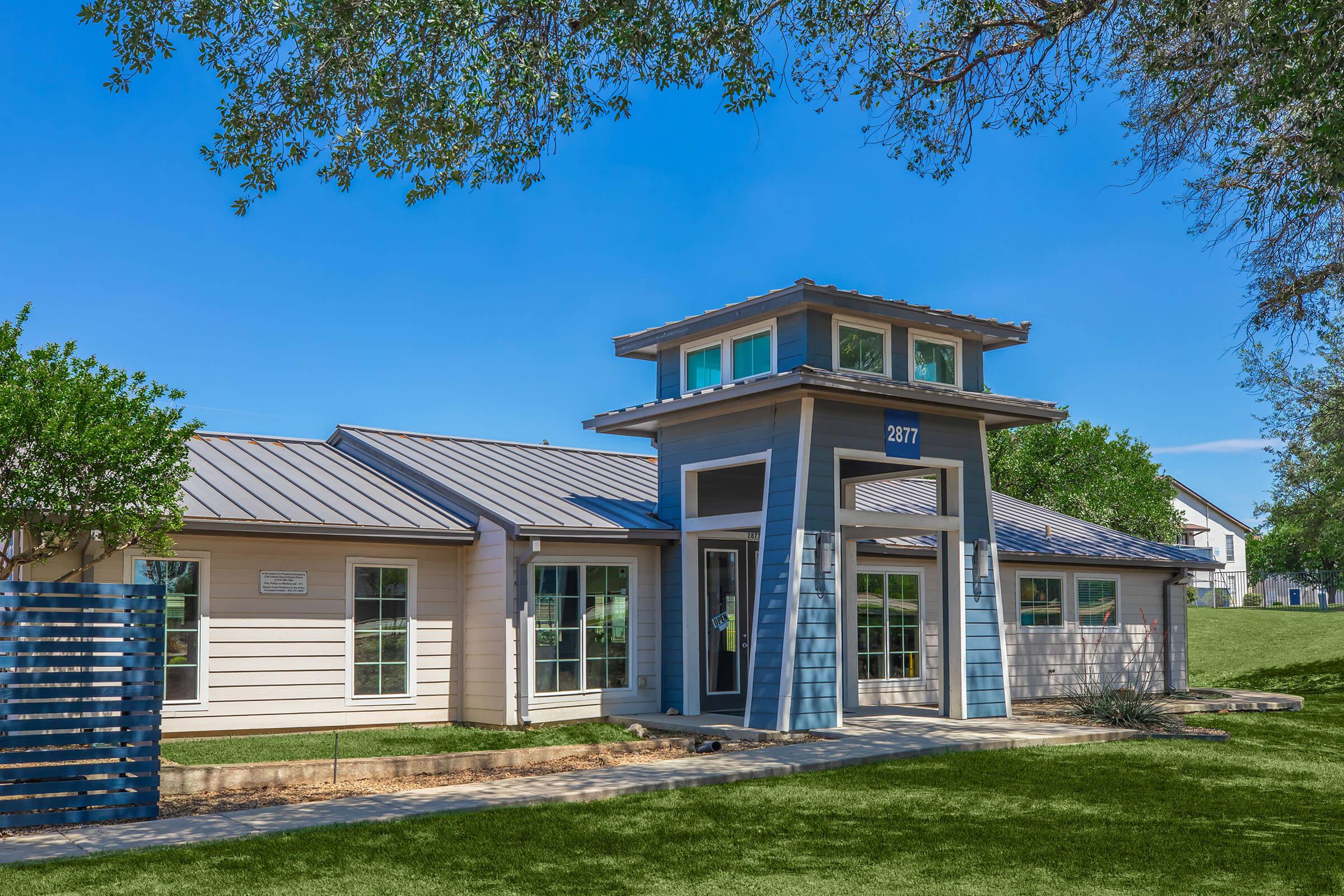A modern single-story building with a light-colored exterior and a distinct tower-like entrance. The structure features large windows and a landscaped green area in front. A clear blue sky enhances the bright and inviting appearance of the facility.