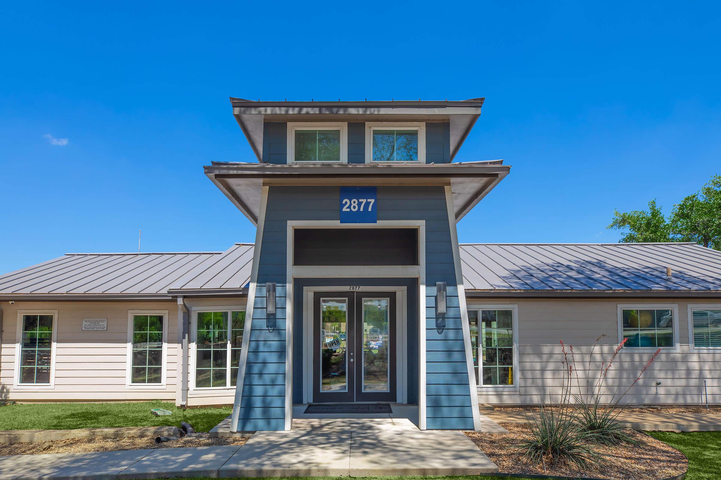 A modern building with a distinctive tower feature, displaying the number "2877" on the front. The structure has a combination of light-colored and blue siding, large windows, and a well-maintained lawn with minimal landscaping. The sky is clear and blue, suggesting a sunny day.